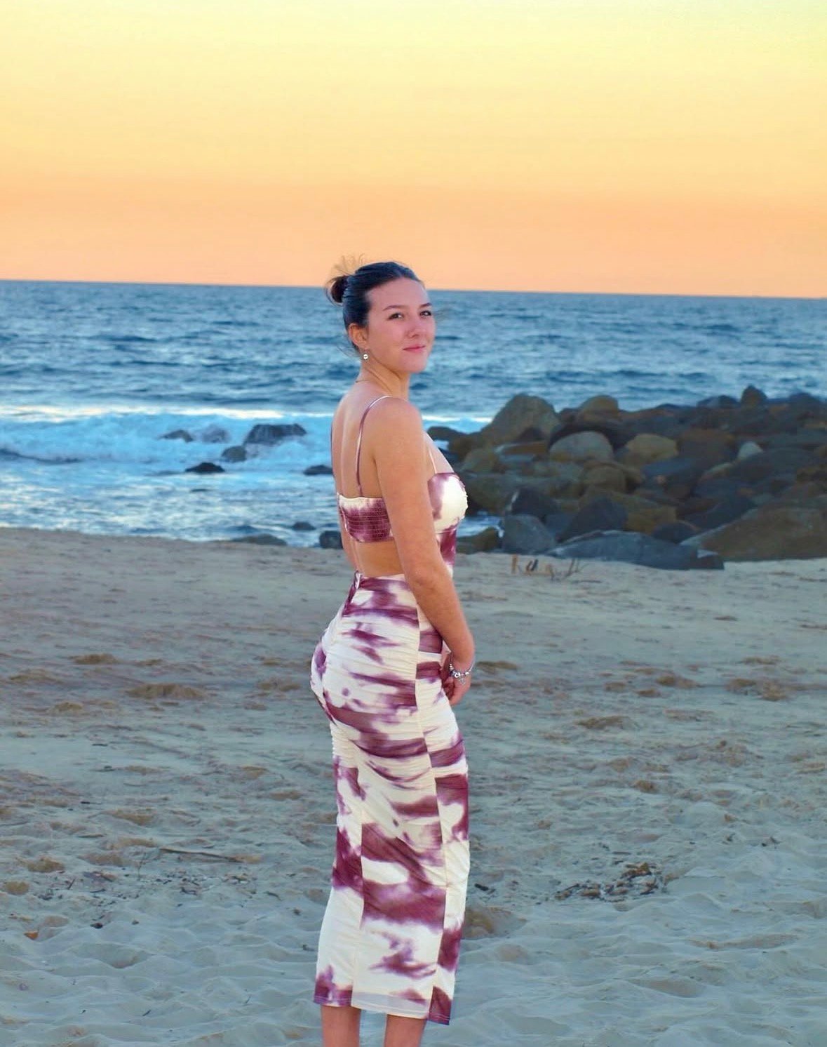 A woman stands on the beach at sunset, wearing a stylish tie-dye outfit. The ocean waves and rocky shoreline are visible in the background.