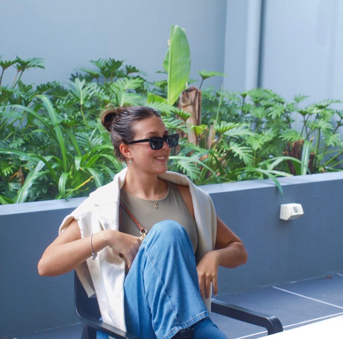 A young woman seated outdoors, wearing sunglasses and a stylish outfit, with lush greenery in the background.