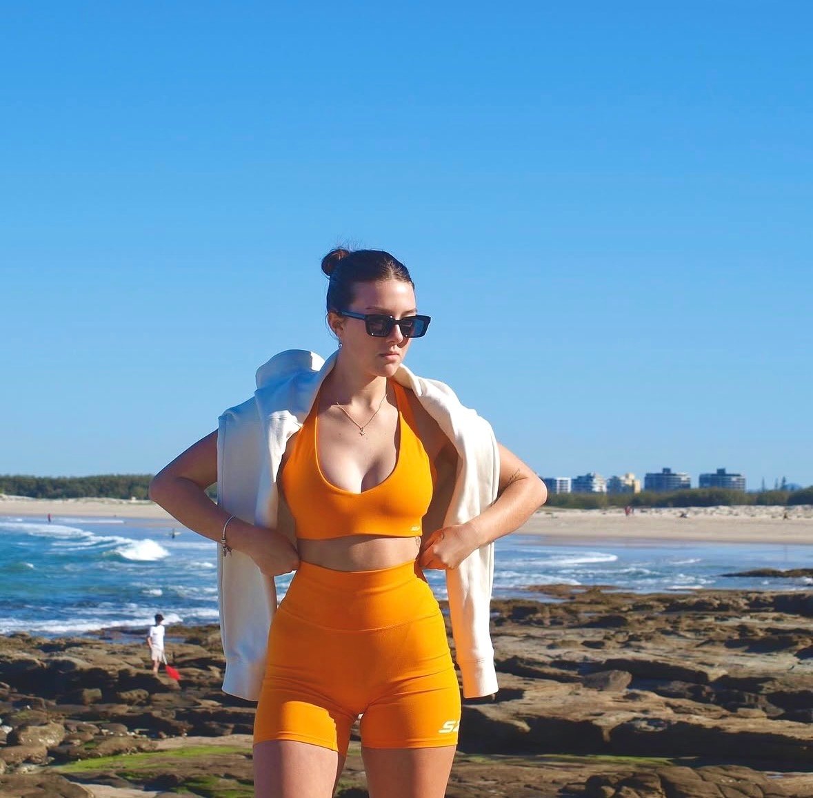A woman in an orange athletic outfit poses confidently on rocky terrain by the beach, with the ocean and city skyline in the background.