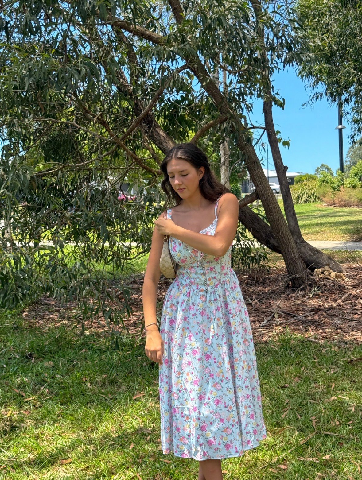 A young woman stands in a sunlit outdoor setting, wearing a floral dress and adjusting her shoulder. Lush greenery surrounds her, creating a serene atmosphere.