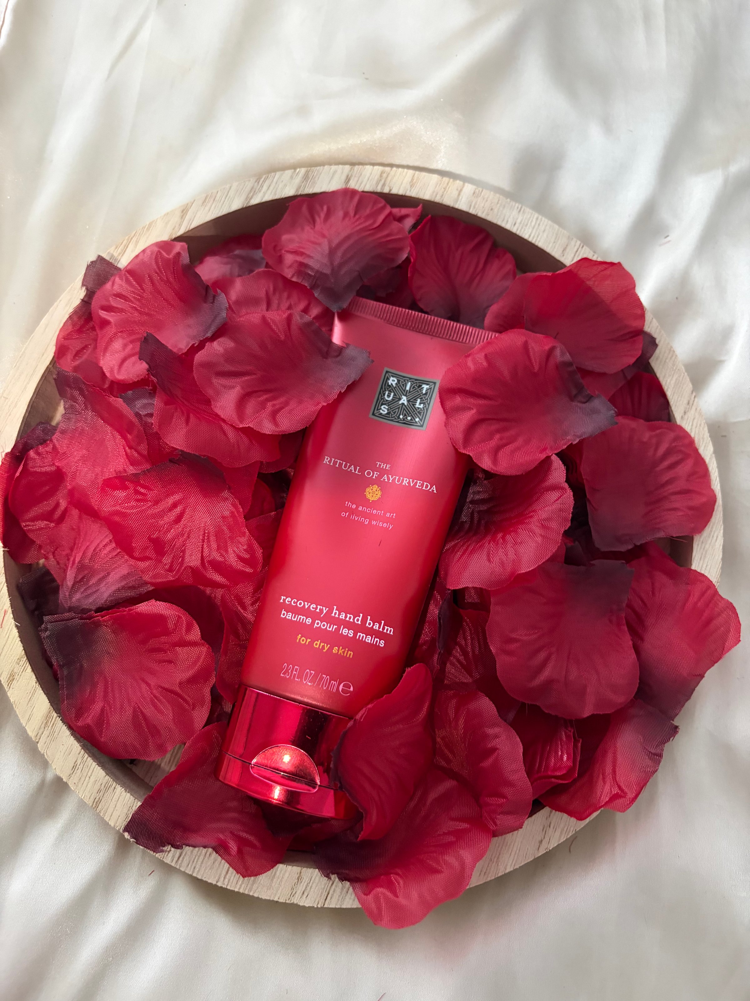 A tube of recovery hand balm surrounded by red rose petals in a wooden bowl.