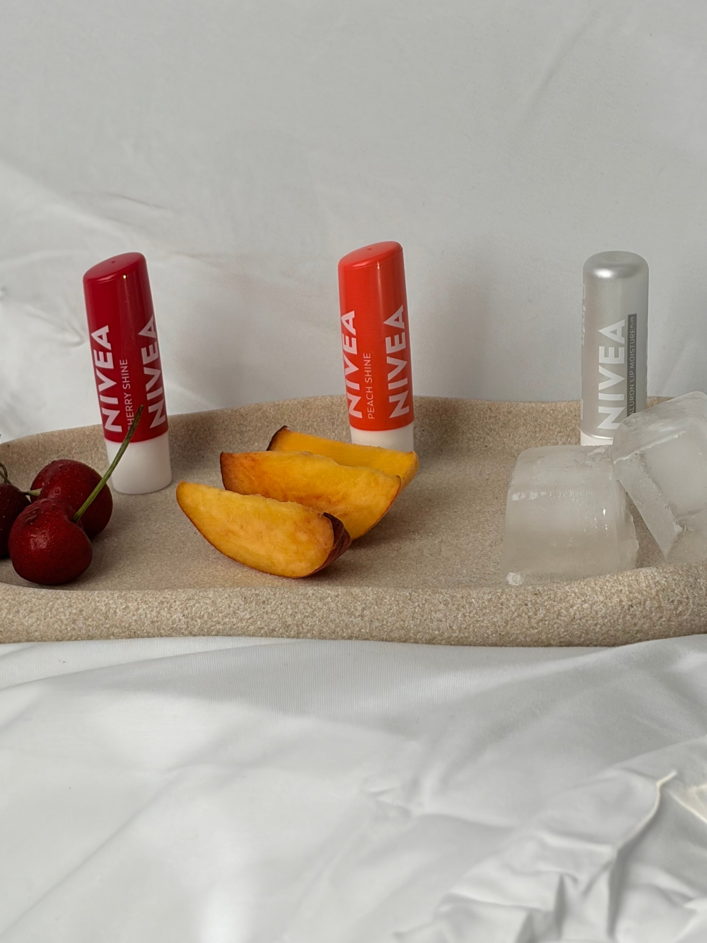 A minimalist arrangement featuring three NIVEA lip balms alongside slices of peach, cherries, and ice cubes on a textured tray.