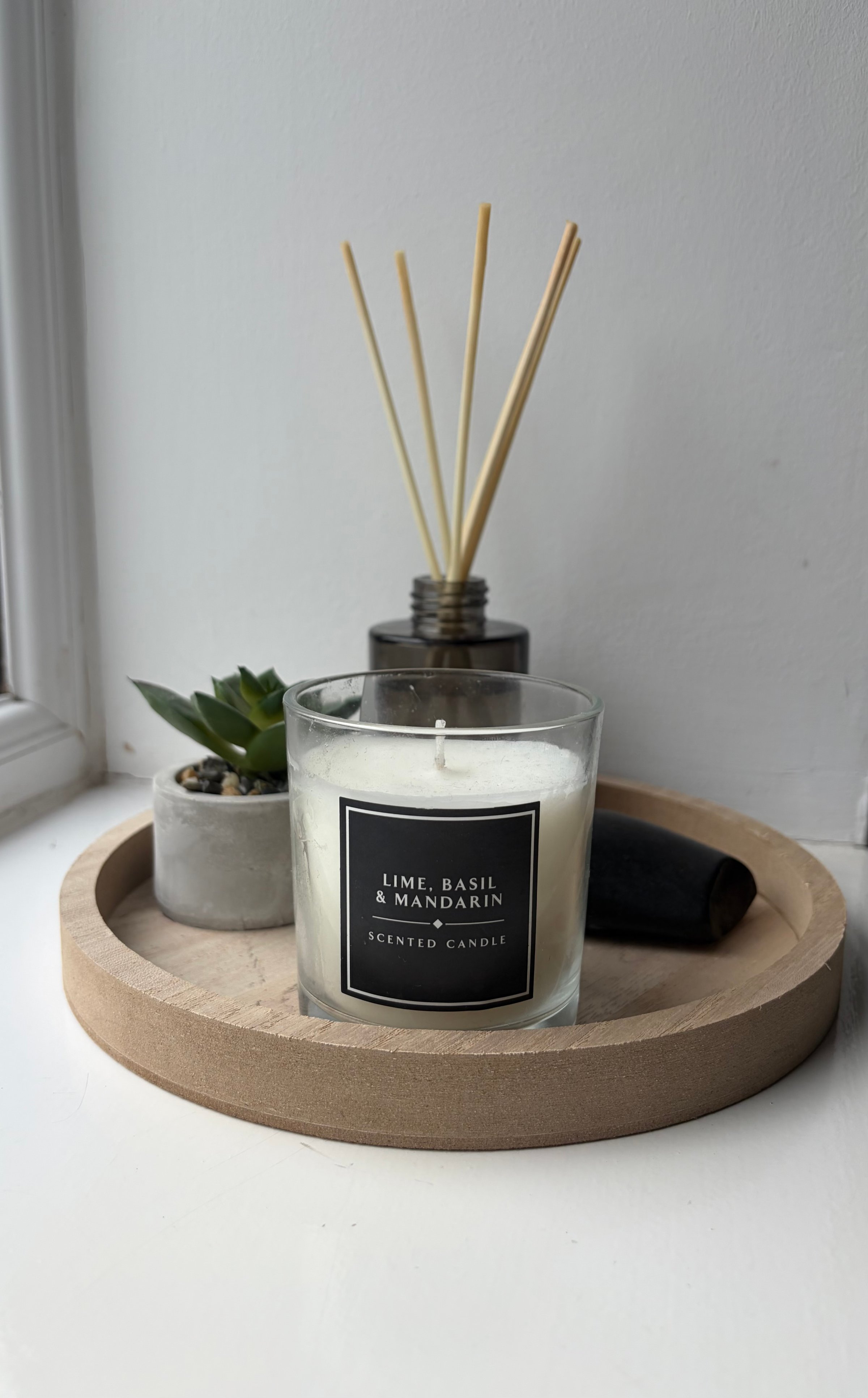 A decorative arrangement featuring a scented candle labeled 'Lime, Basil & Mandarin' on a wooden tray, accompanied by a small succulent plant and a diffuser.