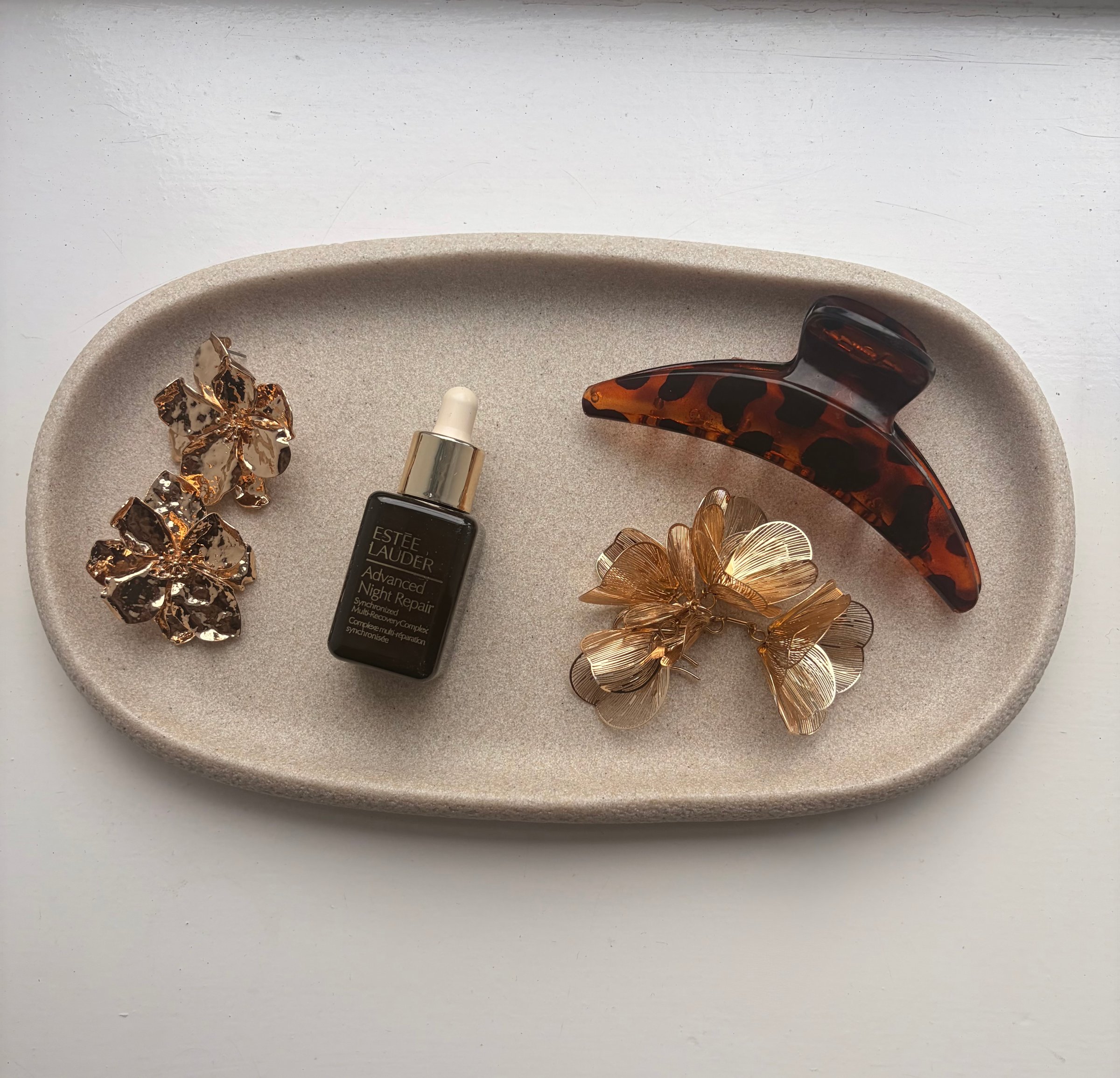 A flat lay of hair accessories and a skincare product arranged on a textured tray. The items include a serum bottle, a tortoiseshell hair clip, and decorative hairpins.