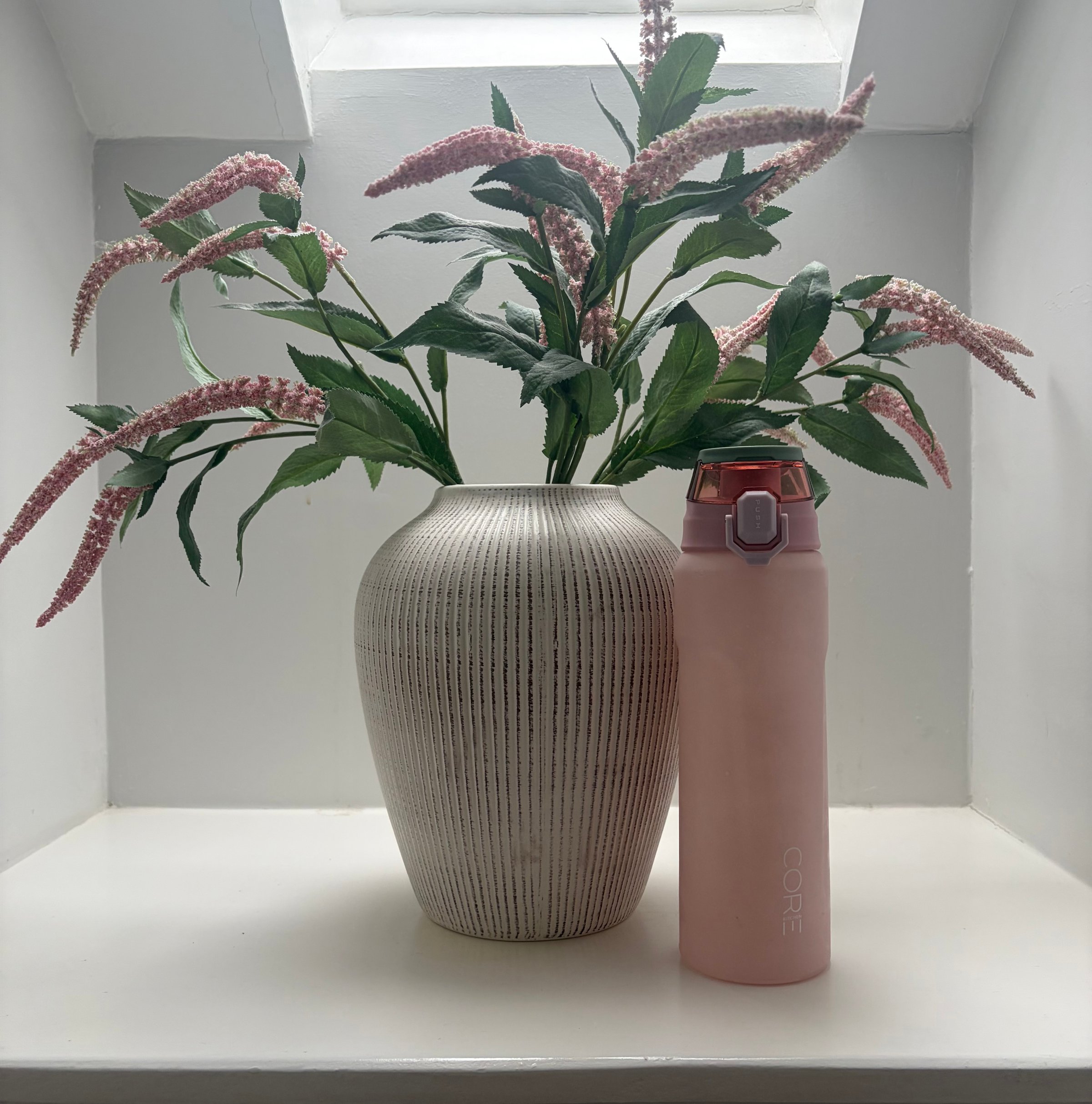 A pink water bottle stands next to a textured white vase filled with pink flowers, placed on a white shelf under a window.