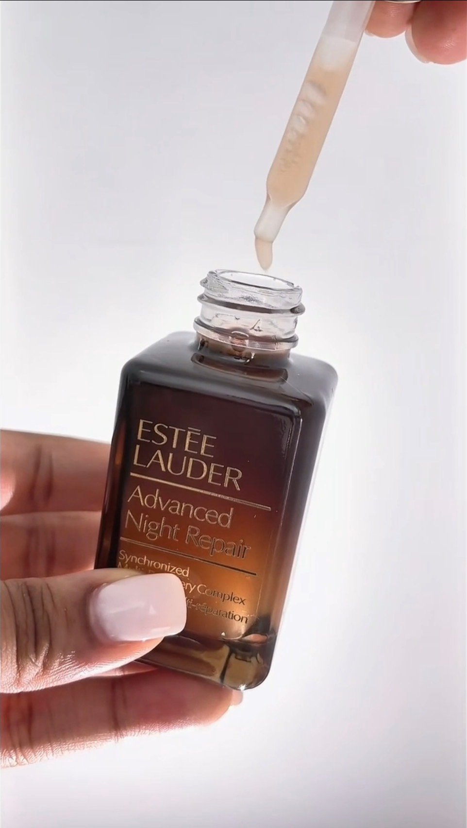 A hand holding a dropper above a bottle of EstΓ©e Lauder Advanced Night Repair serum, with a light background.