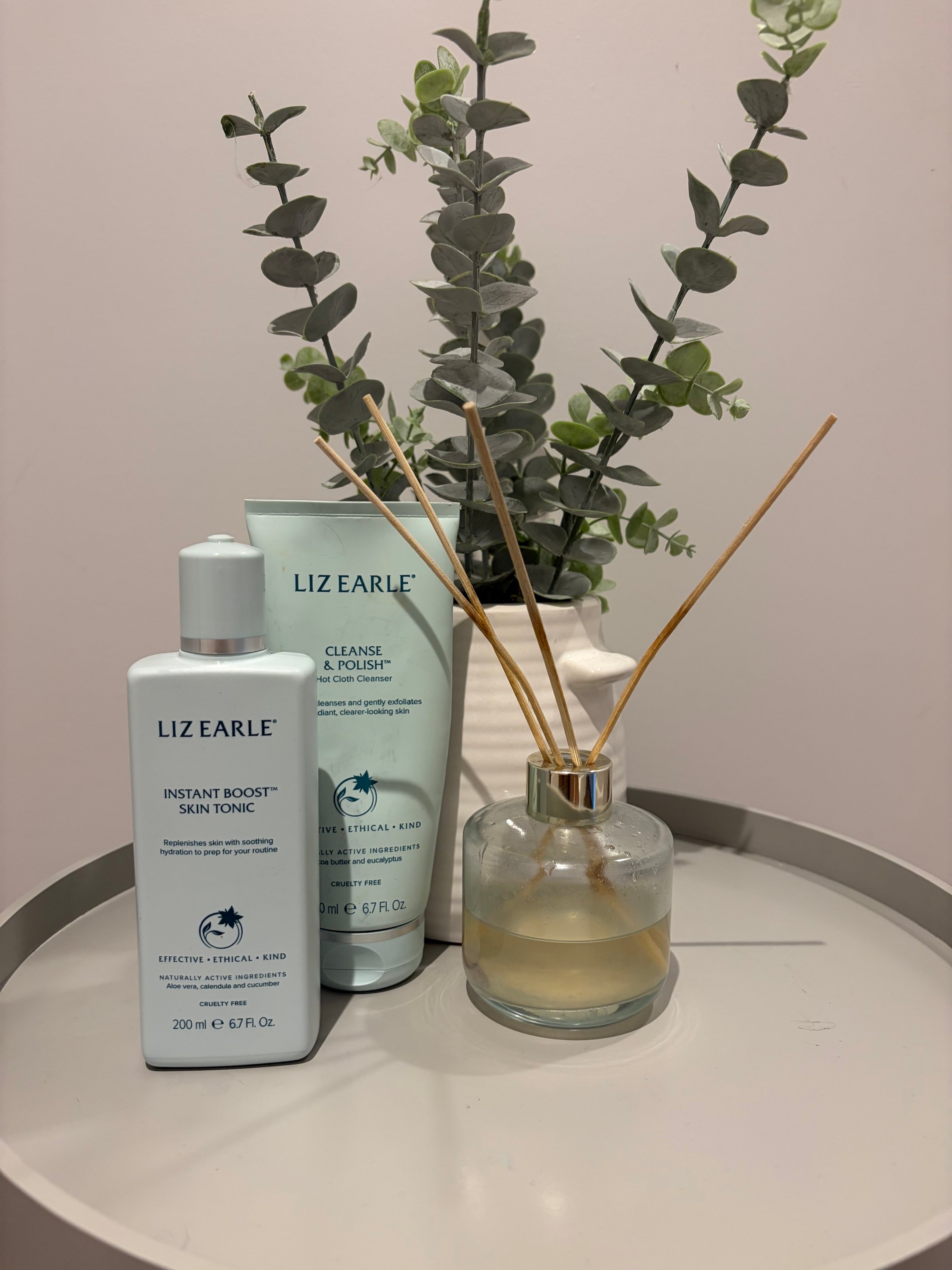 A collection of skincare products from Liz Earle displayed on a round table, accompanied by a decorative plant and a diffuser.