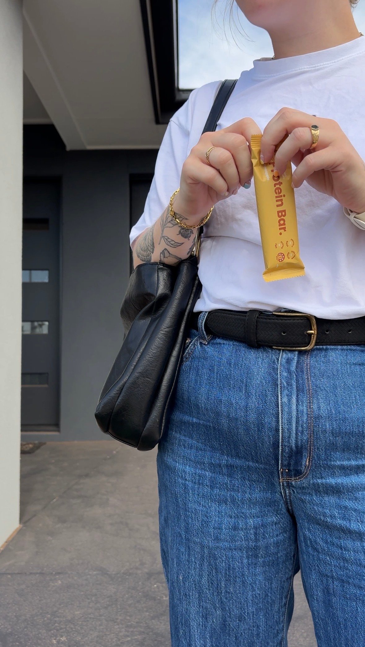 A person holding a protein bar while wearing a casual outfit consisting of a white t-shirt and high-waisted jeans, standing in front of a modern building.