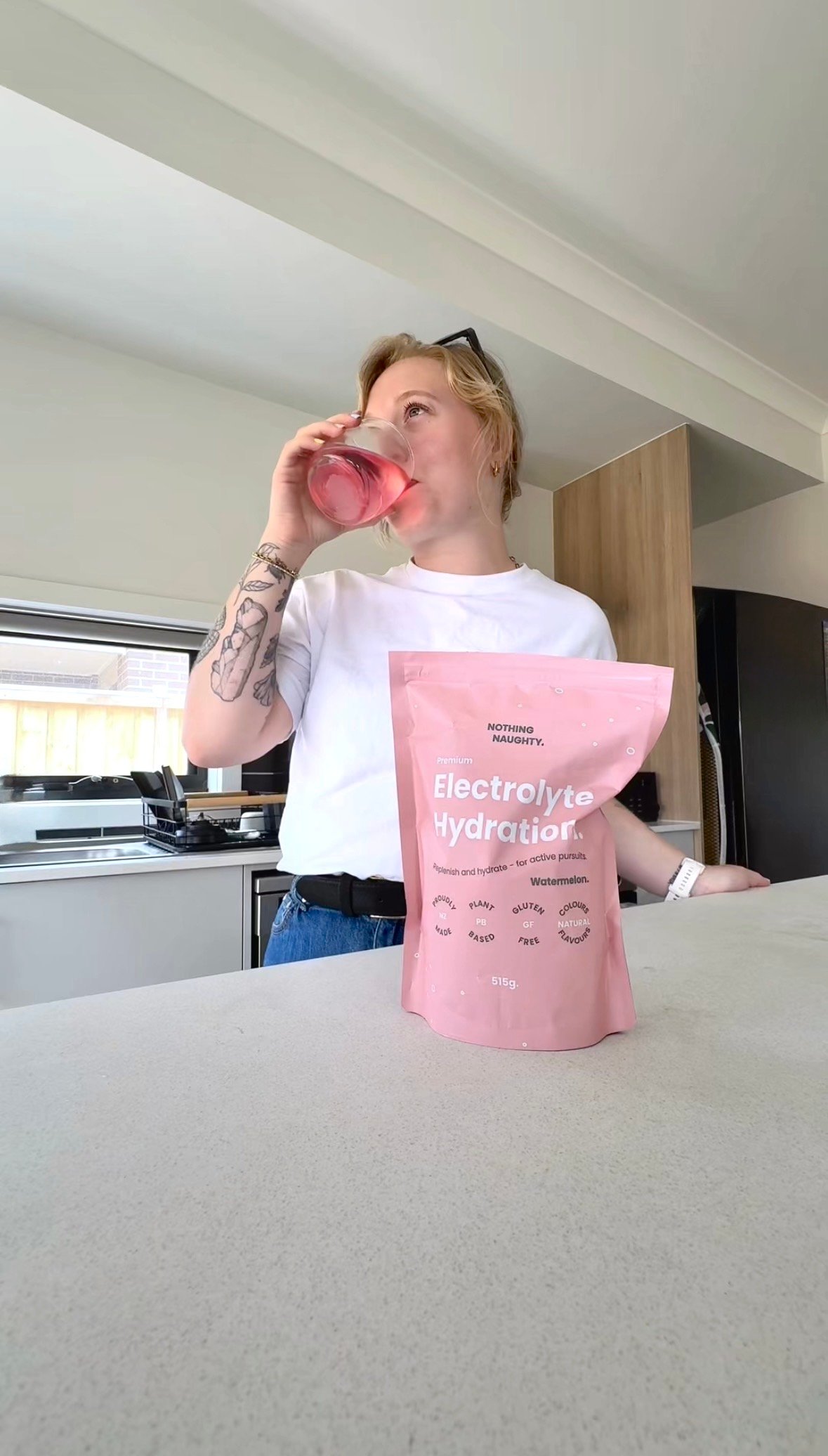 A person with short blonde hair drinks from a glass while standing in a kitchen, with a pink packet of electrolyte hydration mix on the counter.