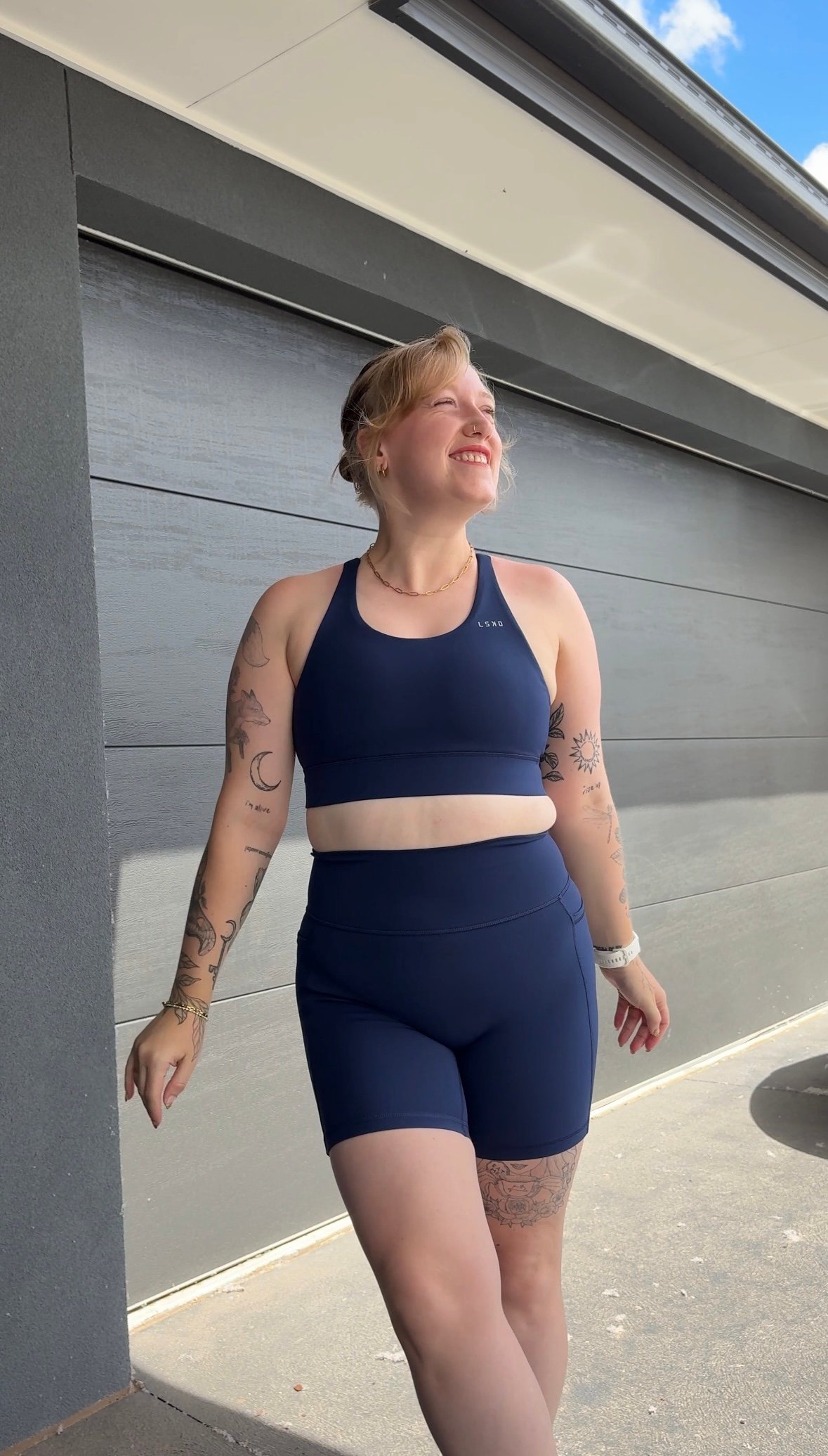 A person wearing a navy blue athletic outfit stands outside, smiling and looking up towards the sky. They have tattoos on their arms and are positioned against a gray wall.