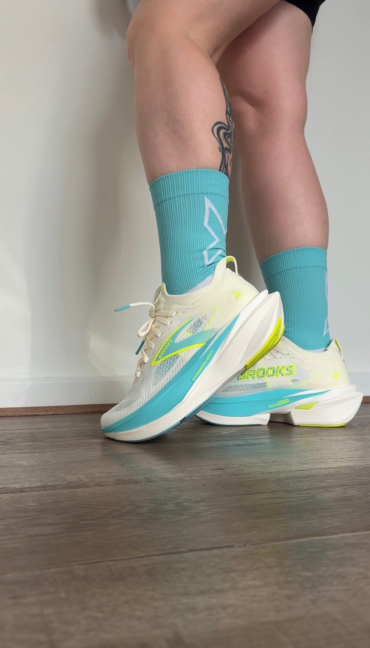 A close-up of a person's legs wearing bright running shoes and teal socks, showcasing a tattoo on the calf.