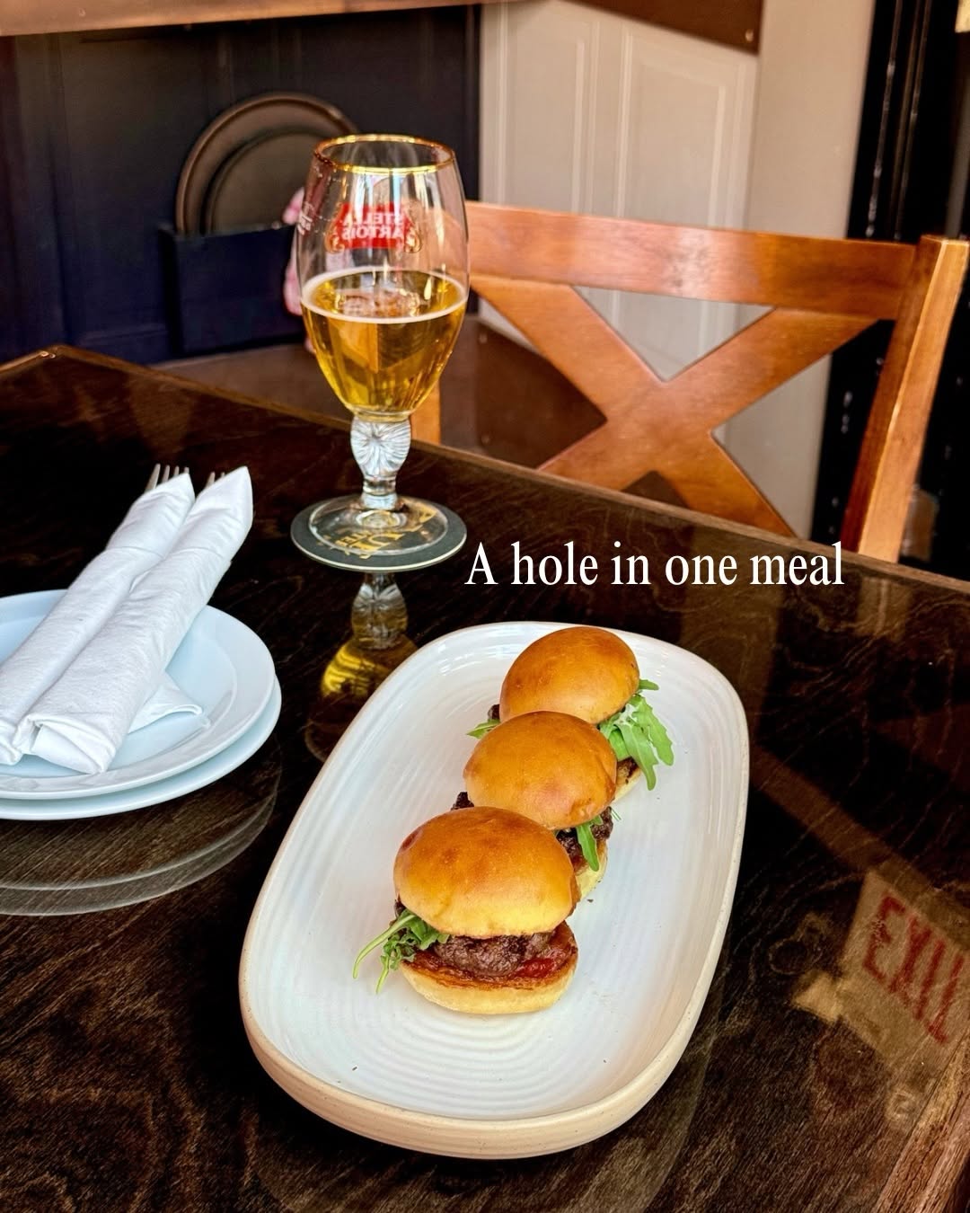 Three mini burgers served on a white plate with arugula, accompanied by a glass of beer on a wooden table.