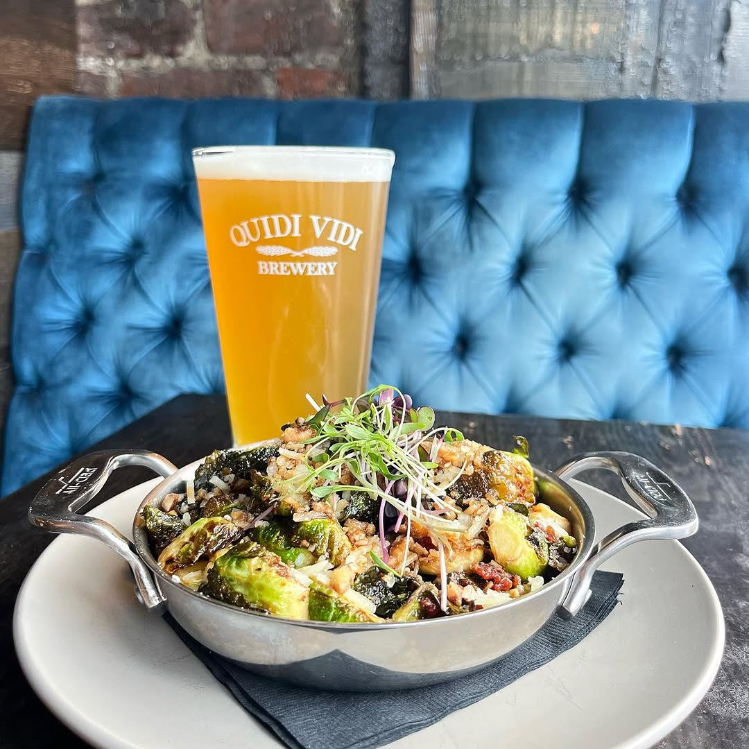 A bowl of Brussels sprouts topped with microgreens sits on a plate next to a glass of beer, against a blue tufted background.