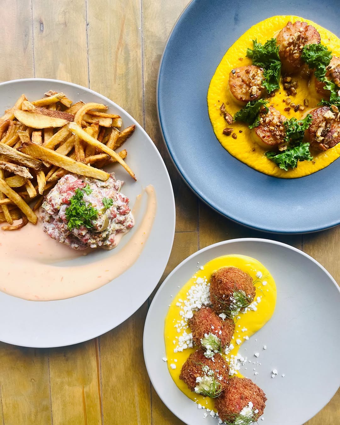 A beautifully arranged plate featuring three different dishes: crispy fries, a seafood salad, and croquettes served with a vibrant yellow sauce.