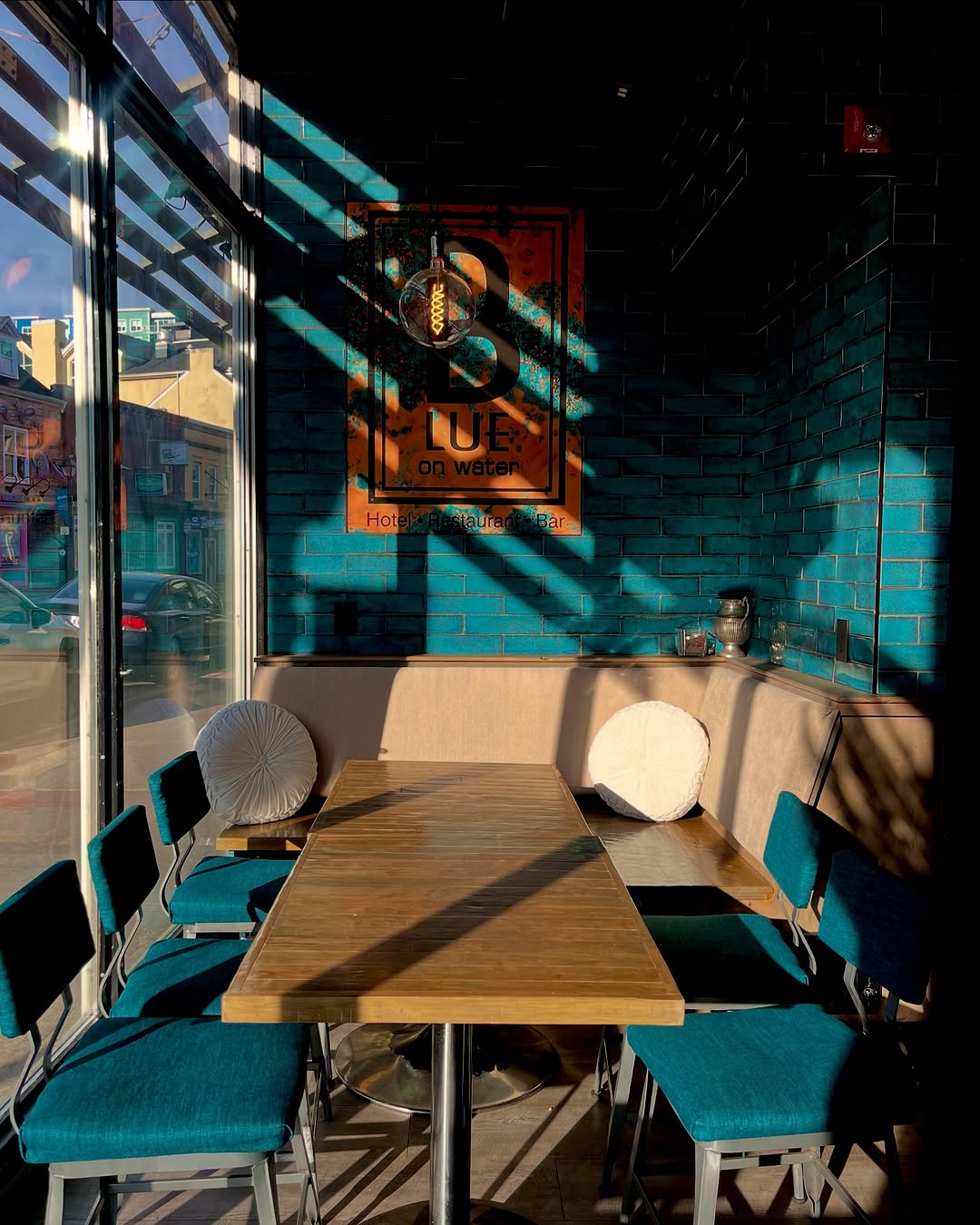 A cozy café interior featuring a wooden table surrounded by teal chairs, with sunlight casting shadows on the blue brick wall.