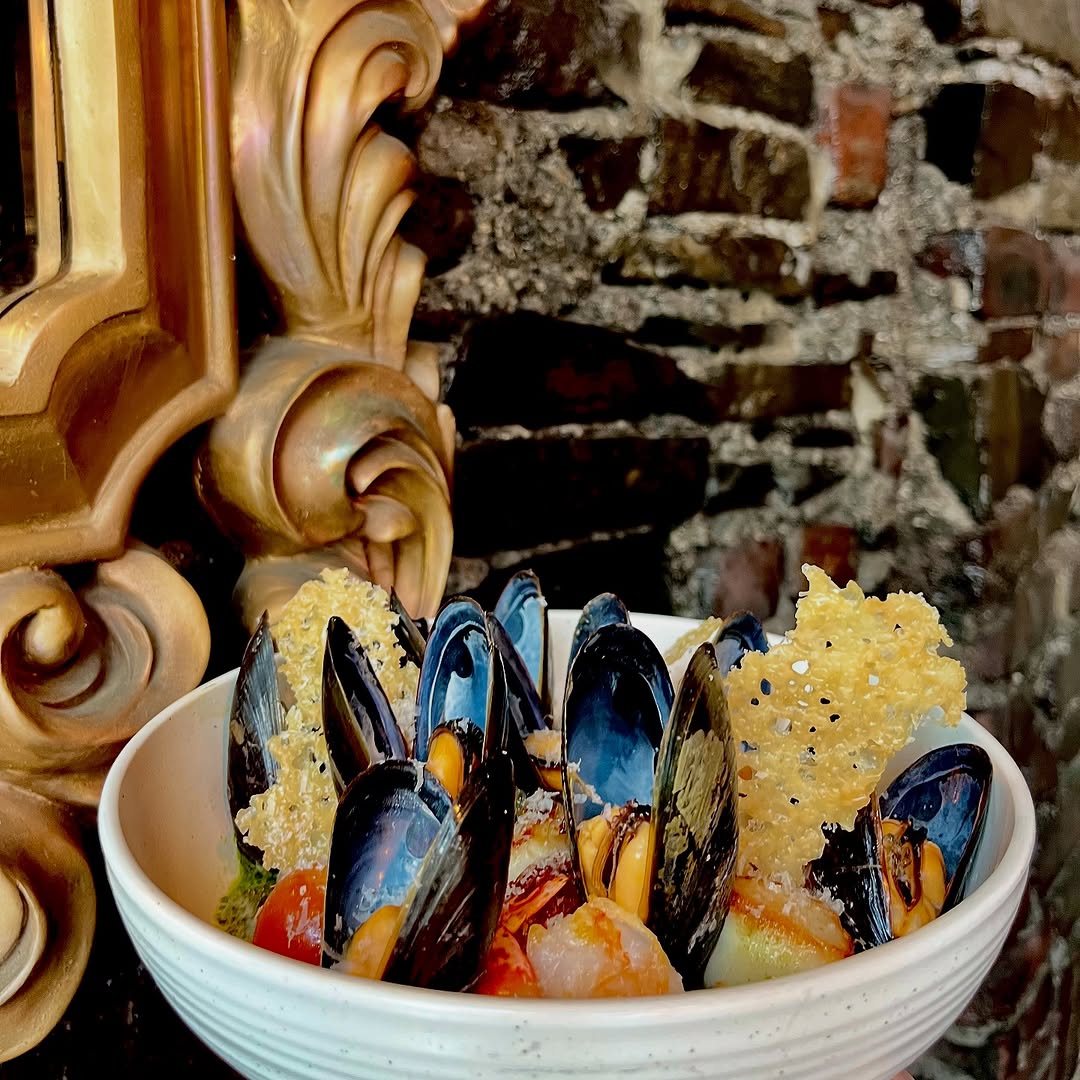A bowl of mussels garnished with crispy cheese and colorful vegetables, set against a textured brick wall.