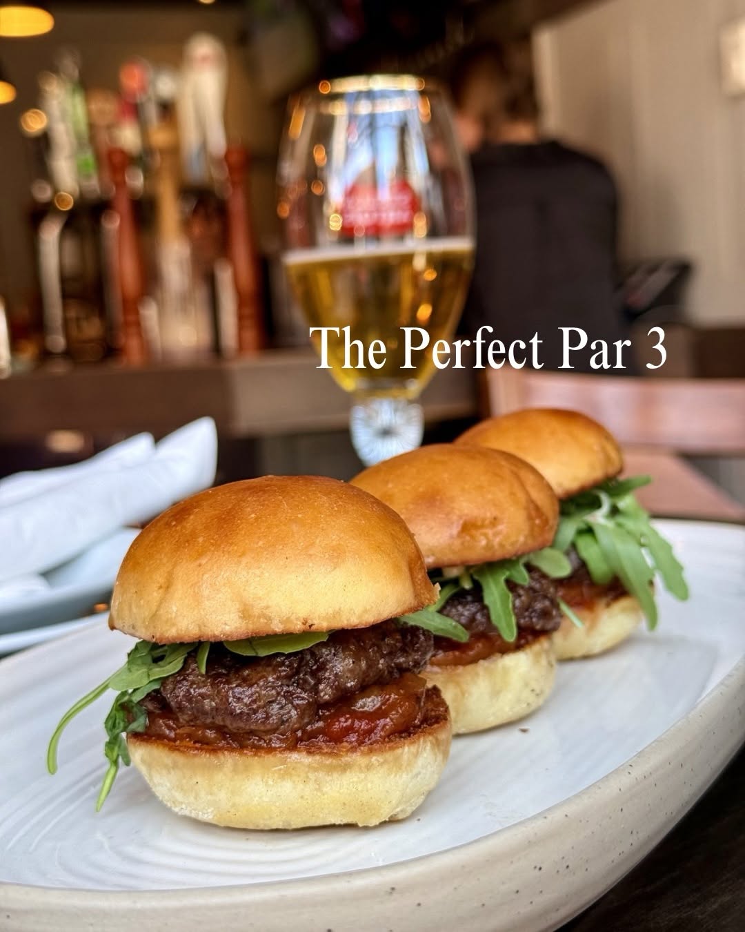 Three mini burgers with arugula served on a white plate, with a glass of beer in the background.