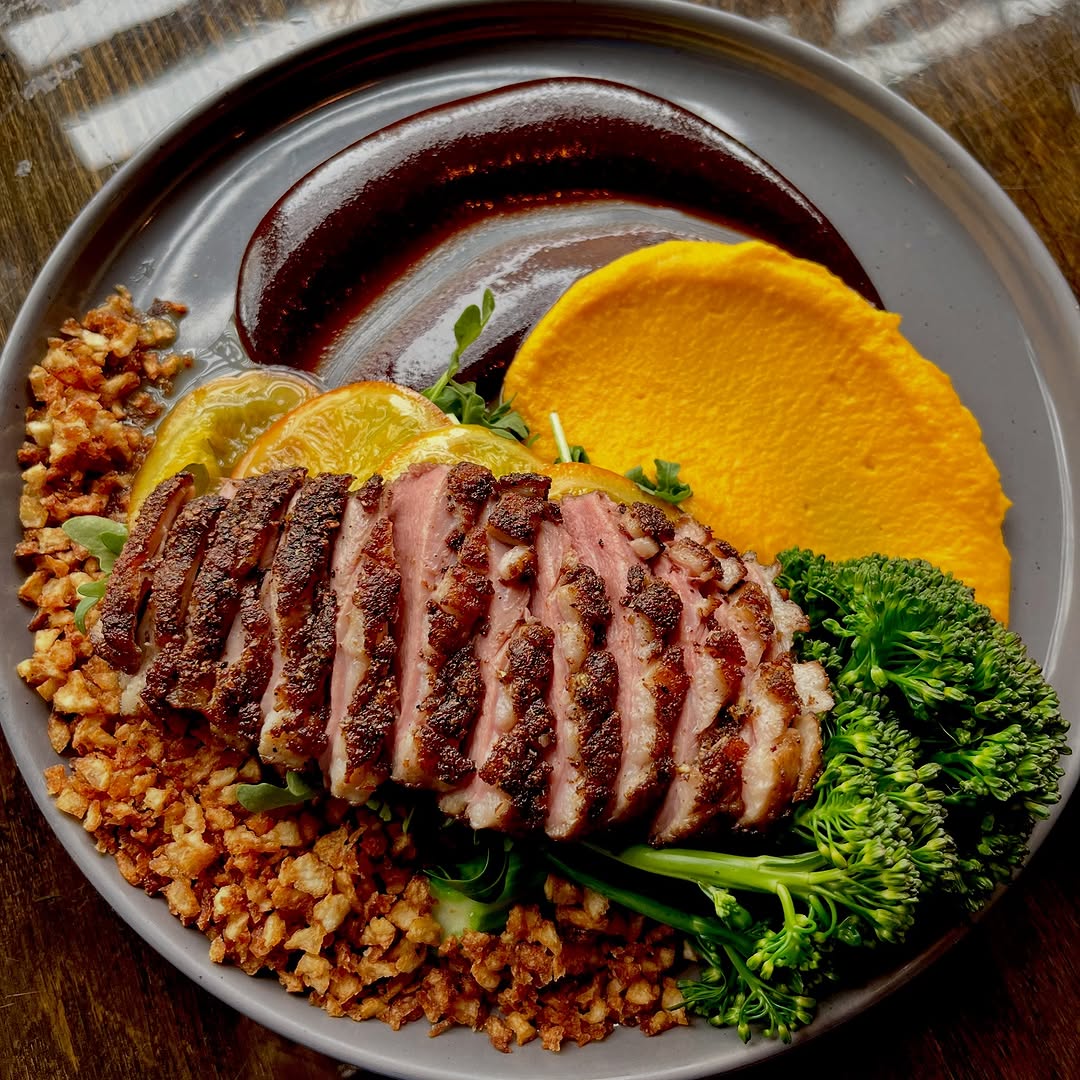 A beautifully plated dish featuring sliced duck breast, accompanied by a vibrant orange puree, broccolini, and a dark sauce, garnished with lemon slices and grains.