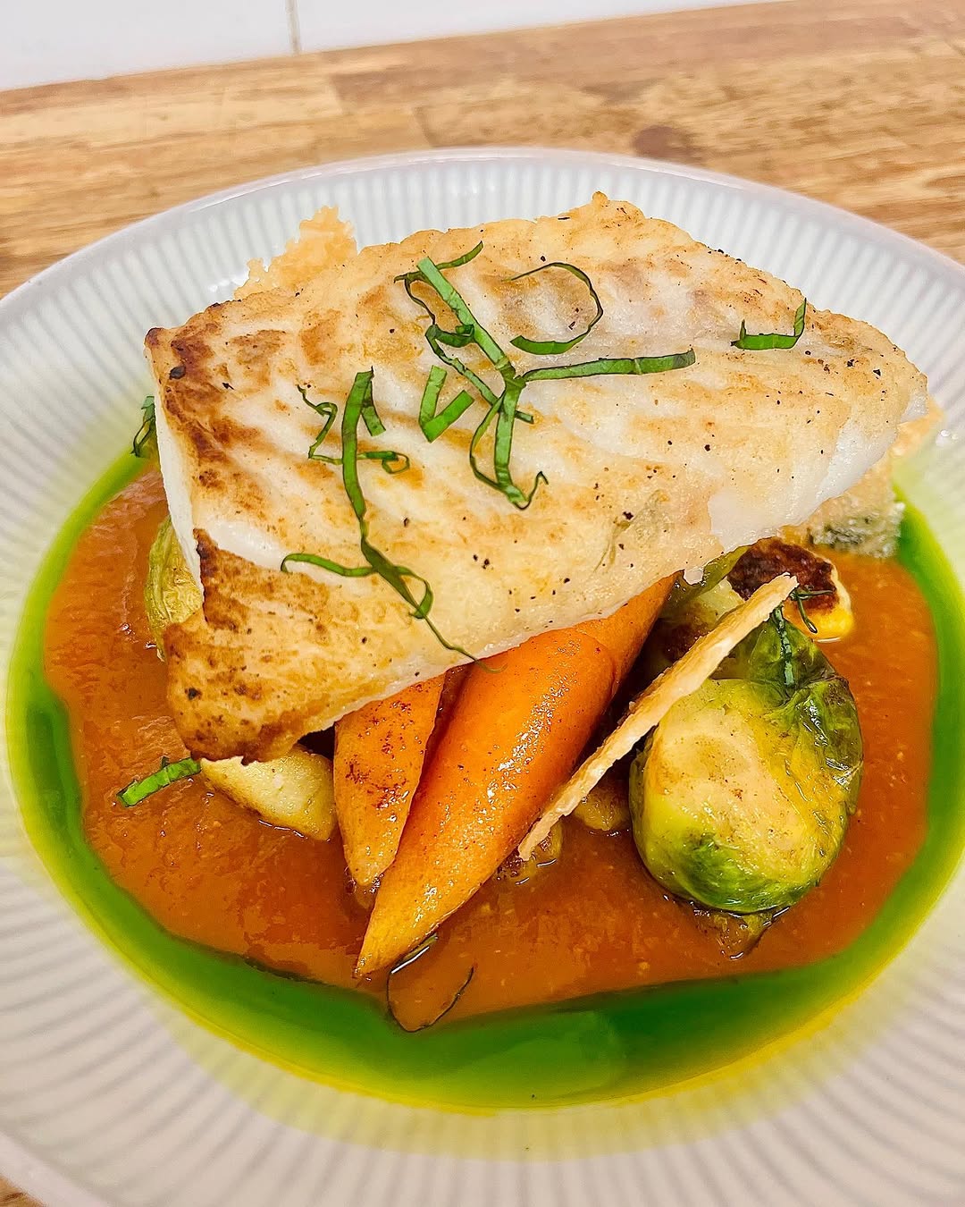 A beautifully plated dish featuring a piece of fish atop a colorful arrangement of roasted vegetables, including carrots and Brussels sprouts, drizzled with a vibrant green sauce.