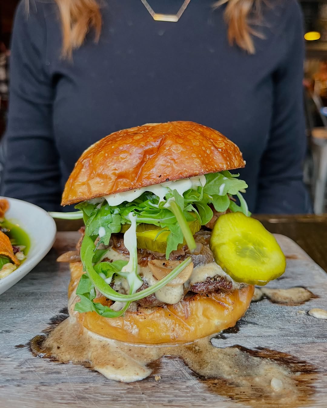A gourmet burger topped with fresh greens, pickles, and creamy sauce, served on a wooden platter.