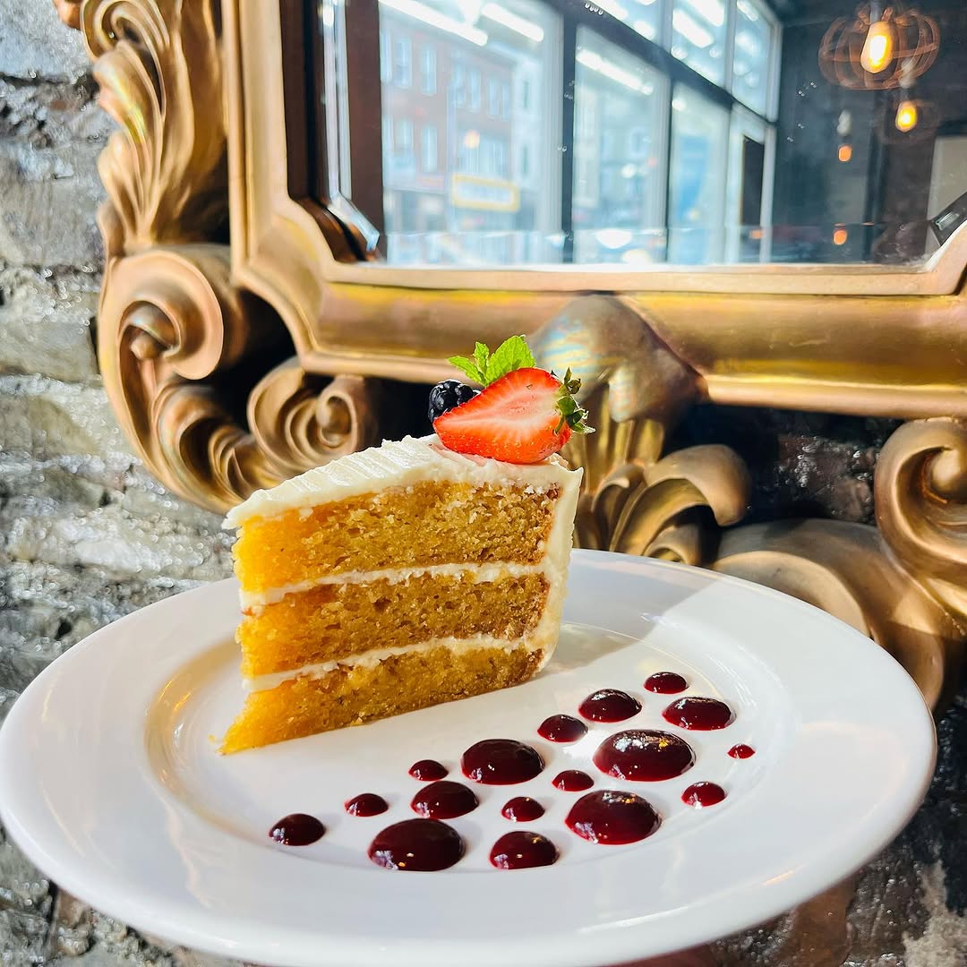 A slice of layered cake topped with frosting, garnished with a strawberry and a blueberry, served on a white plate with a drizzle of berry sauce, set against an ornate golden mirror.