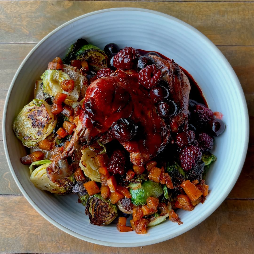 A beautifully arranged plate featuring roasted duck leg topped with a berry sauce, accompanied by Brussels sprouts and a medley of diced vegetables.