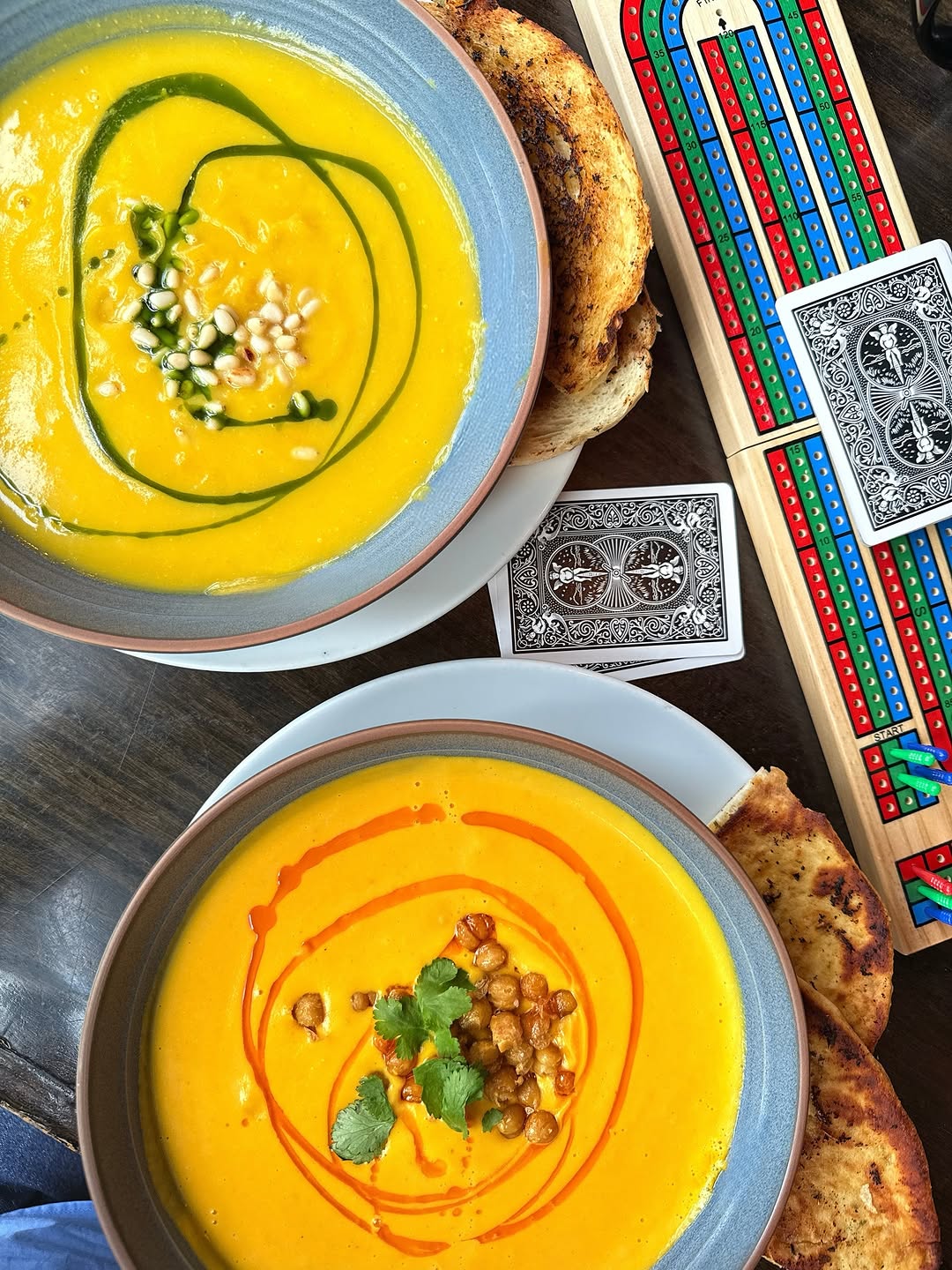 Two bowls of vibrant yellow soup garnished with herbs and spices, accompanied by toasted bread and playing cards on a wooden table.