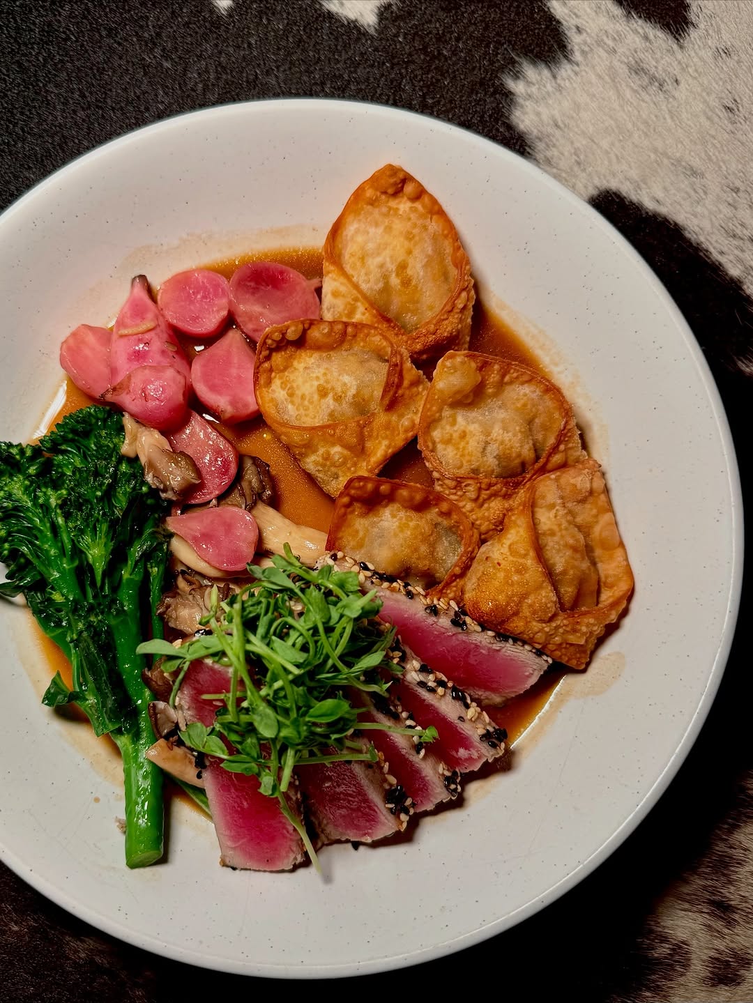 A beautifully arranged plate featuring seared tuna, crispy dumplings, vibrant radishes, and sautéed greens in a savory broth.