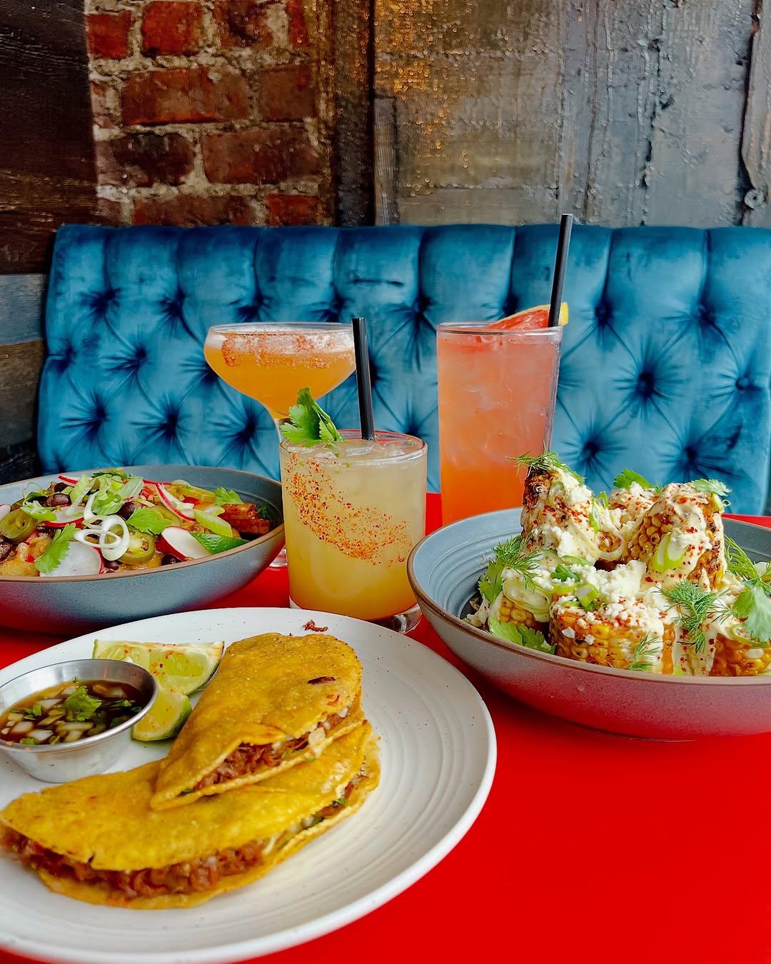 A vibrant table setting featuring a variety of colorful dishes and drinks, including tacos, a salad, and cocktails, against a rustic backdrop.