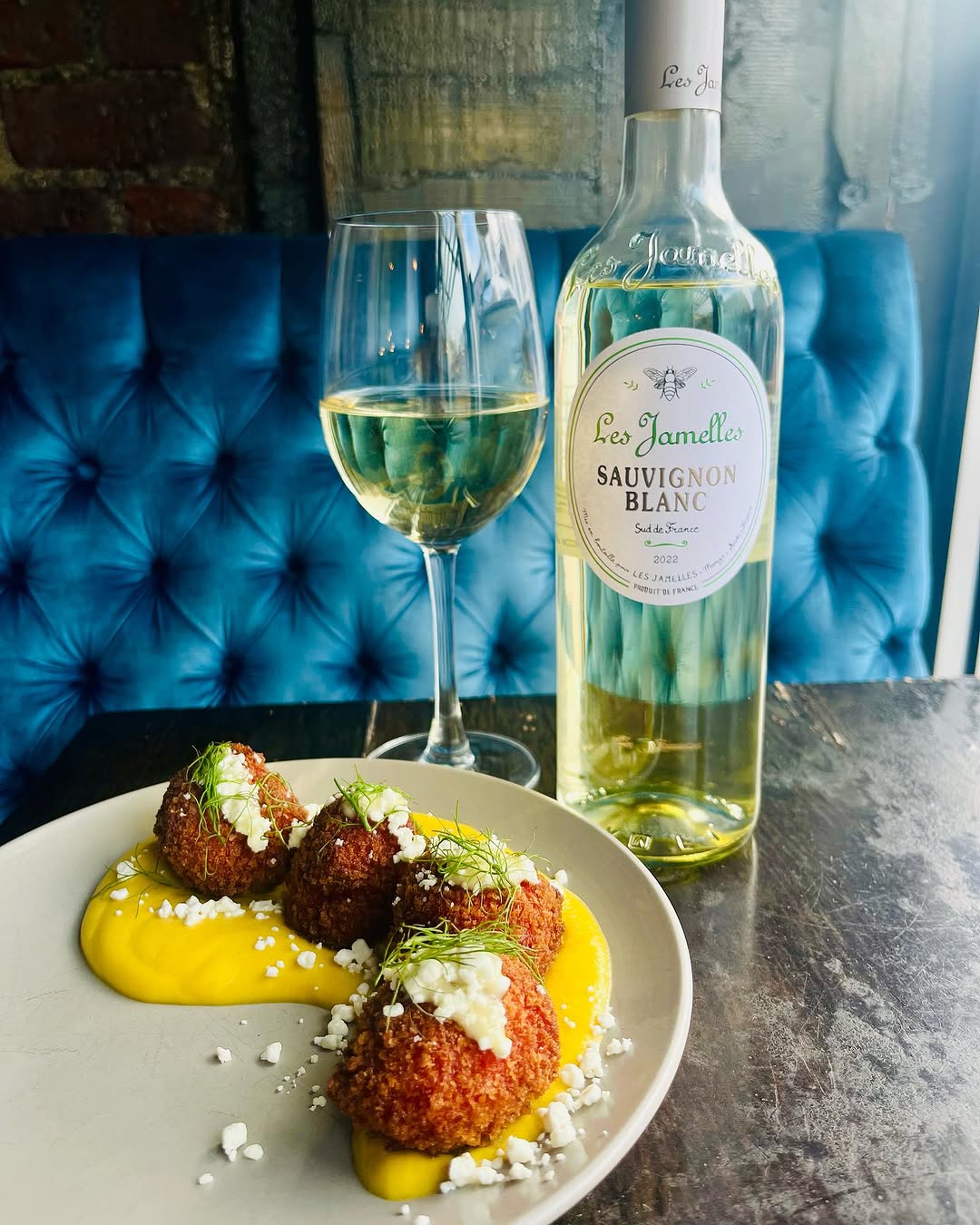 A plate of golden-brown croquettes served with a vibrant yellow sauce, accompanied by a glass of Sauvignon Blanc and a bottle of wine in the background.