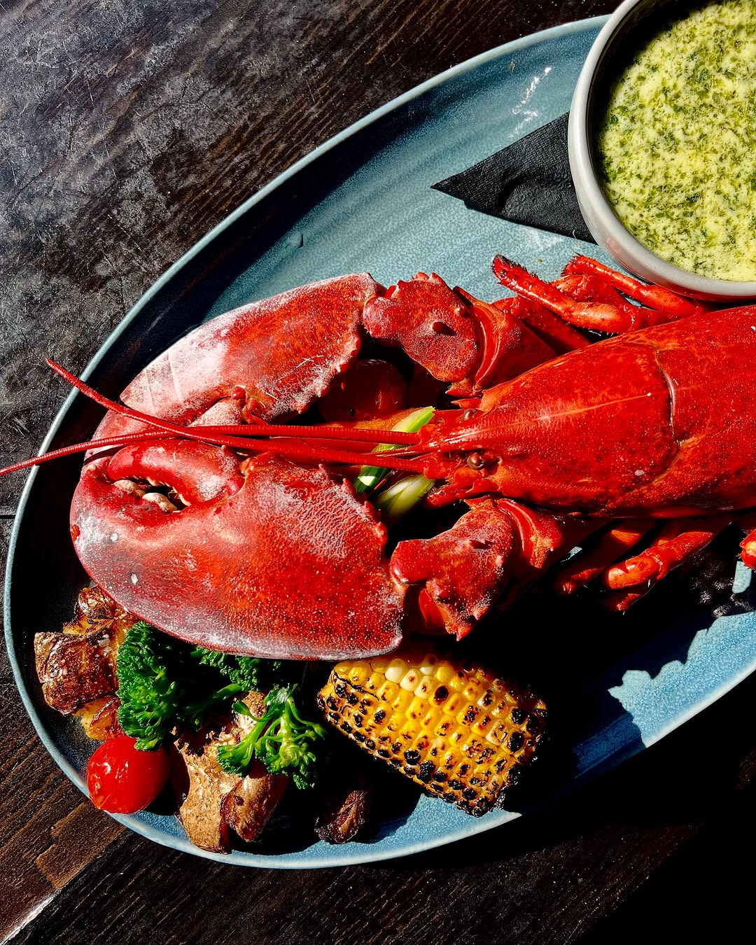 A vibrant plate featuring a whole red lobster alongside grilled vegetables and corn, complemented by a small bowl of green sauce.