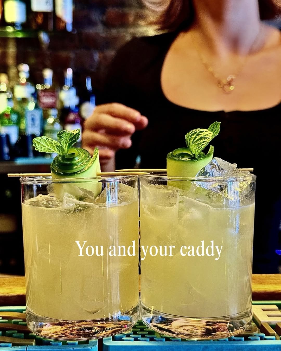 Two cocktails garnished with cucumber and mint, placed on a bar counter with a blurred background of bottles.