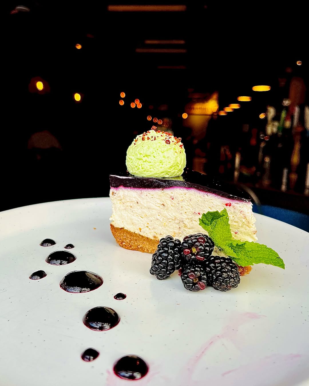 A slice of cheesecake topped with a green mint-flavored dollop and surrounded by blackberries and berry sauce on a white plate.