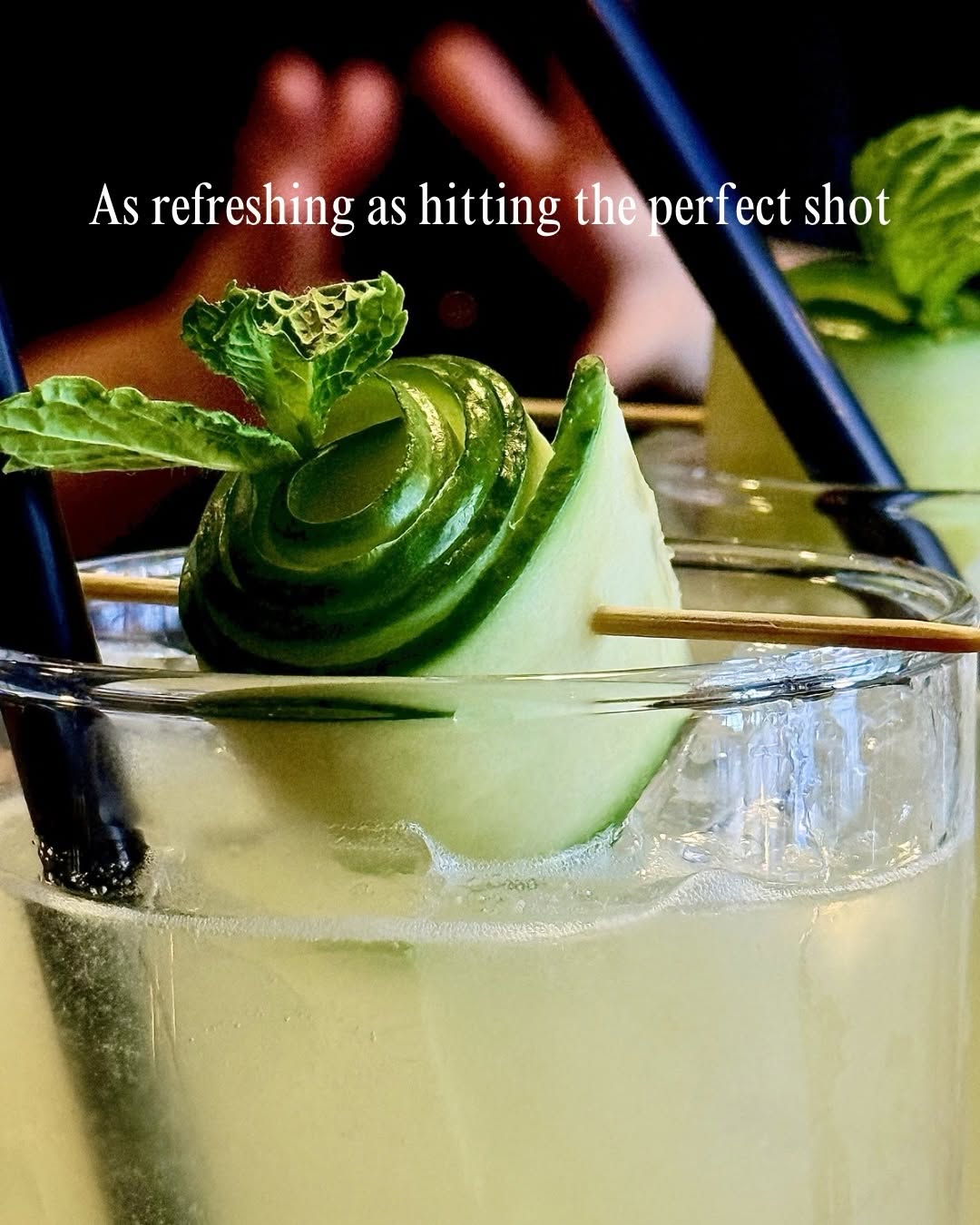 A refreshing cocktail garnished with a spiral of cucumber and mint leaves, served in a clear glass with straws.