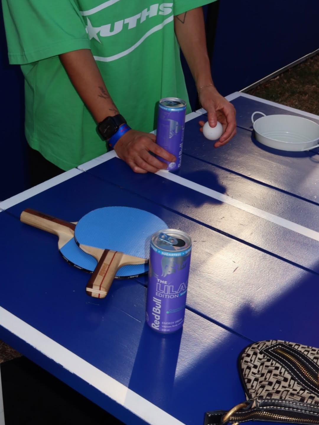 A person holding a ping pong ball and paddle, with two cans of Red Bull positioned on a blue table.