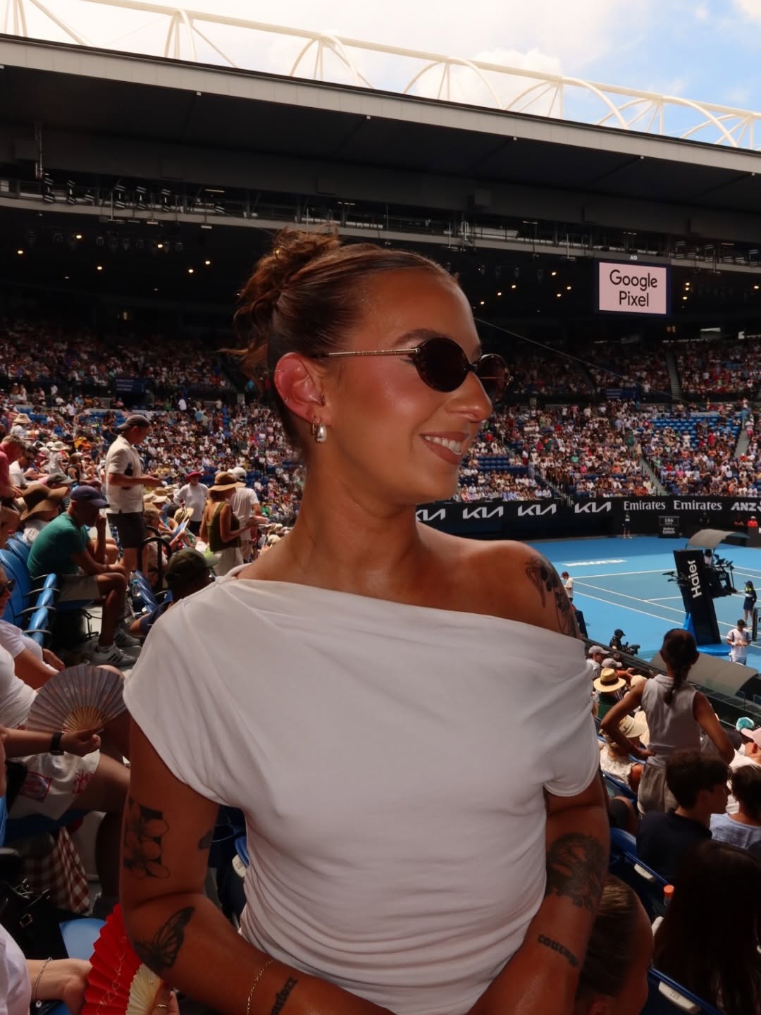 A woman with sunglasses smiles while sitting in a stadium filled with spectators during a tennis match.