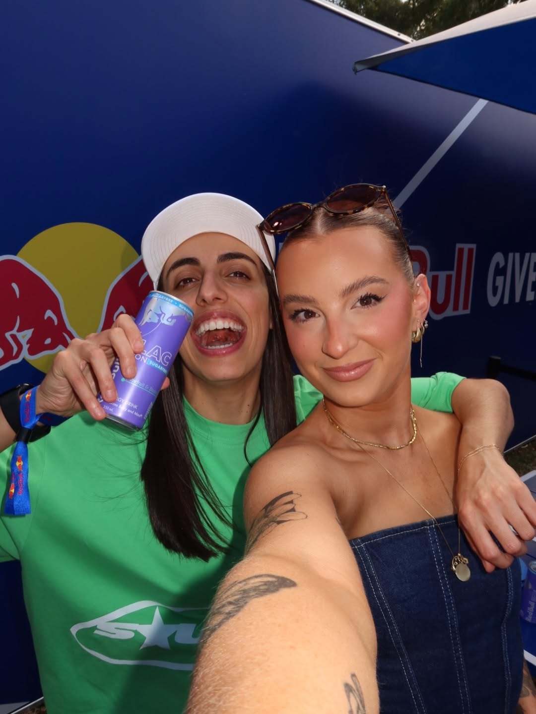 Two women smiling and posing for a selfie at an outdoor event, one holding a beverage can.