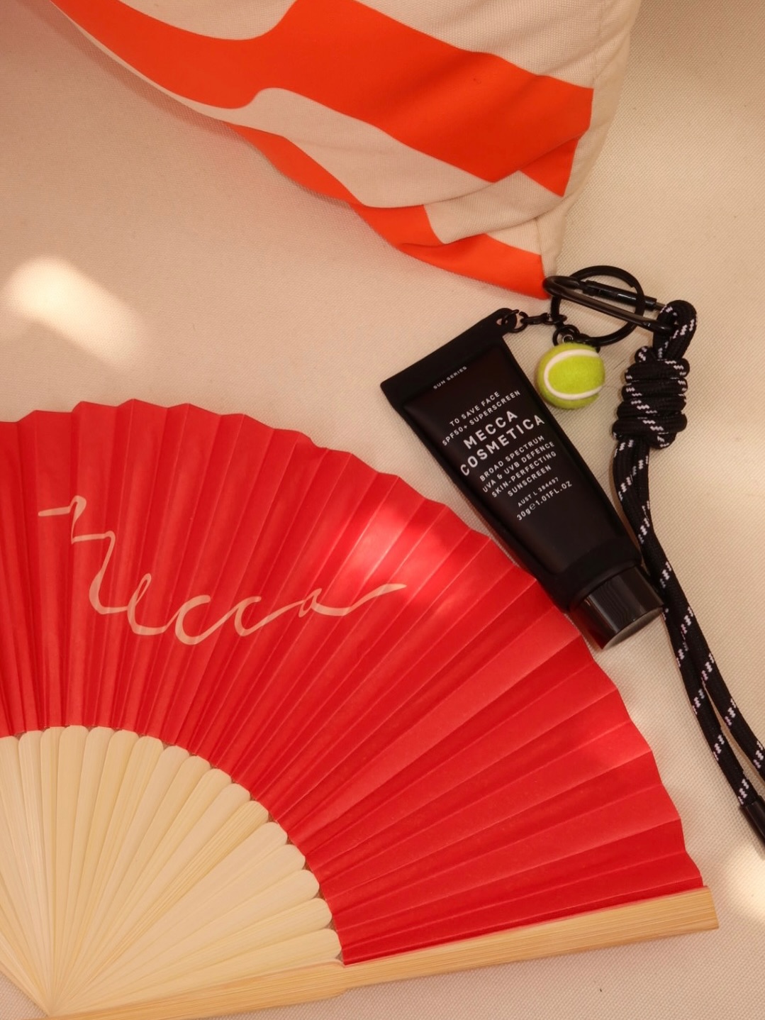 A vibrant red fan and a tube of Mecca Cosmetica skincare product are arranged on a textured surface, complemented by a small green accessory.