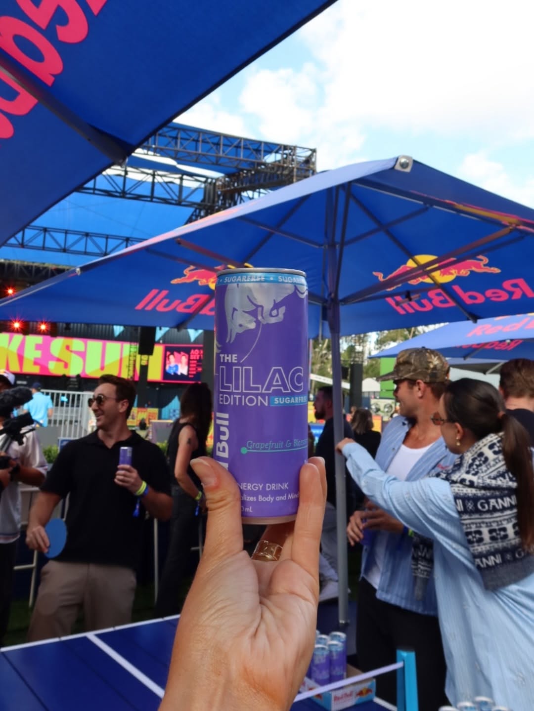 A hand holding a can of Lilac Edition energy drink at an outdoor event, with people socializing in the background under blue canopies.