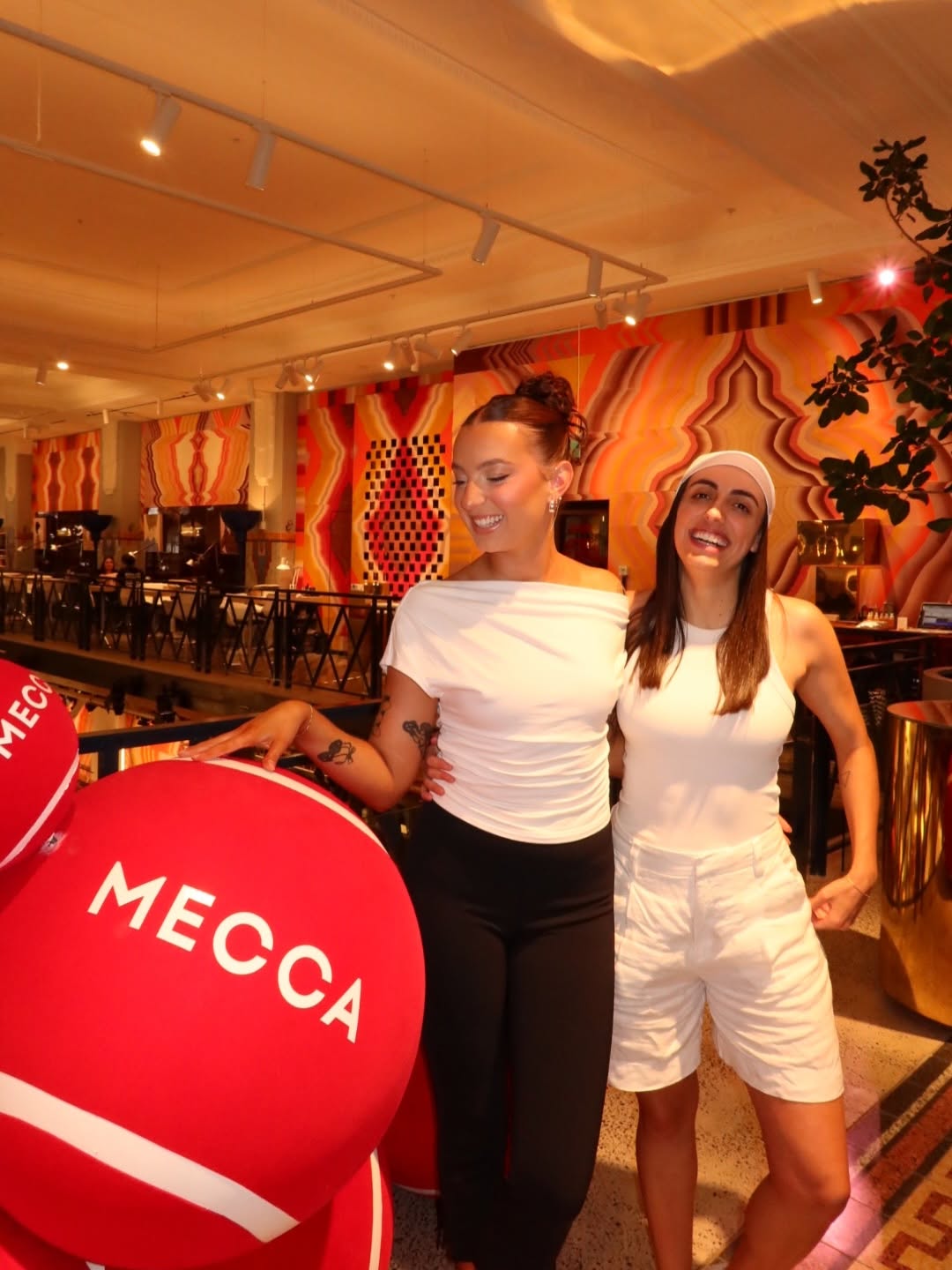 Two women pose playfully in a vibrant store setting, surrounded by large red promotional balls with 'MECCA' branding.