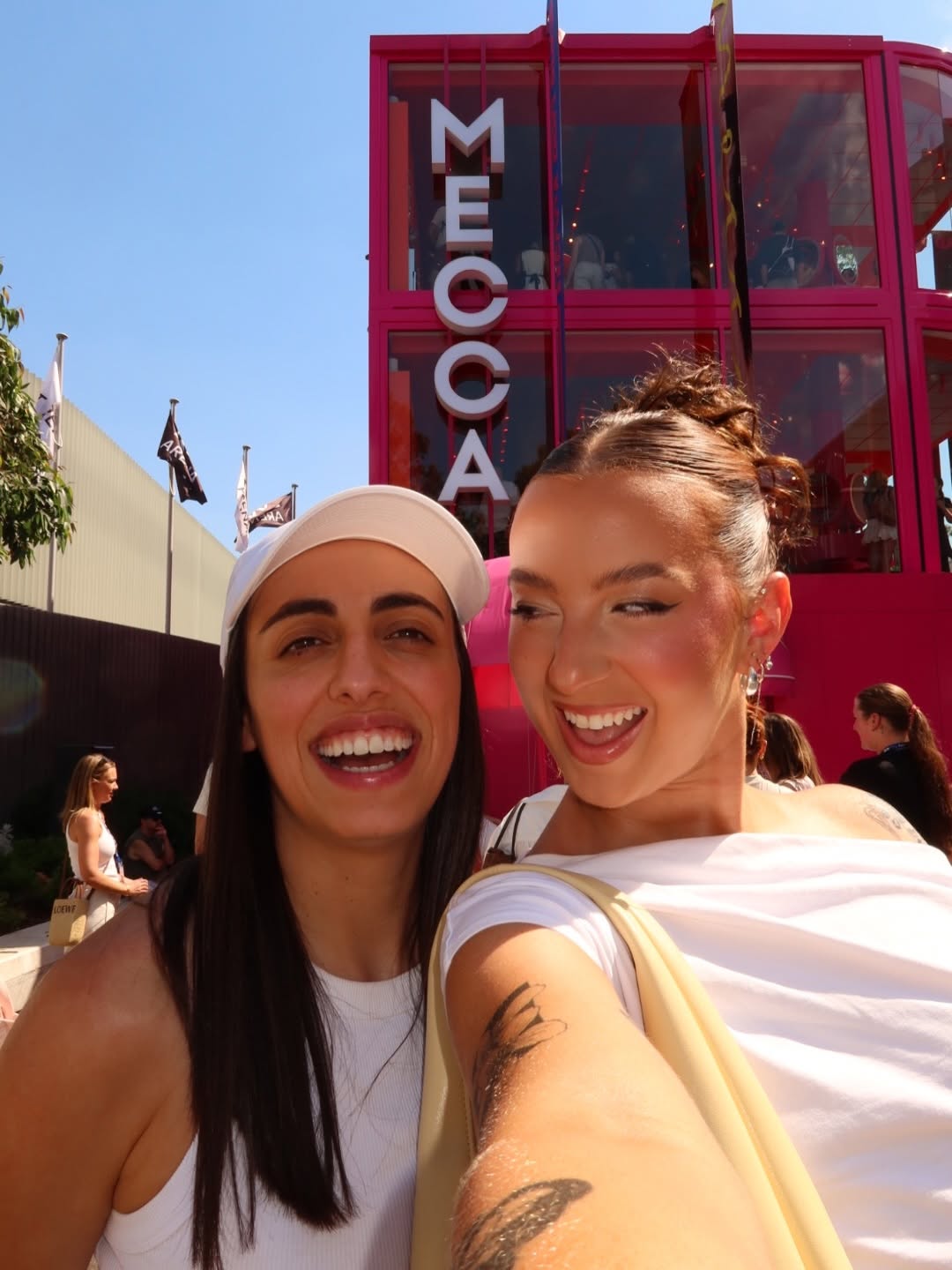 Two women smiling at the camera in front of a vibrant pink building with the word 'MECCA' prominently displayed.