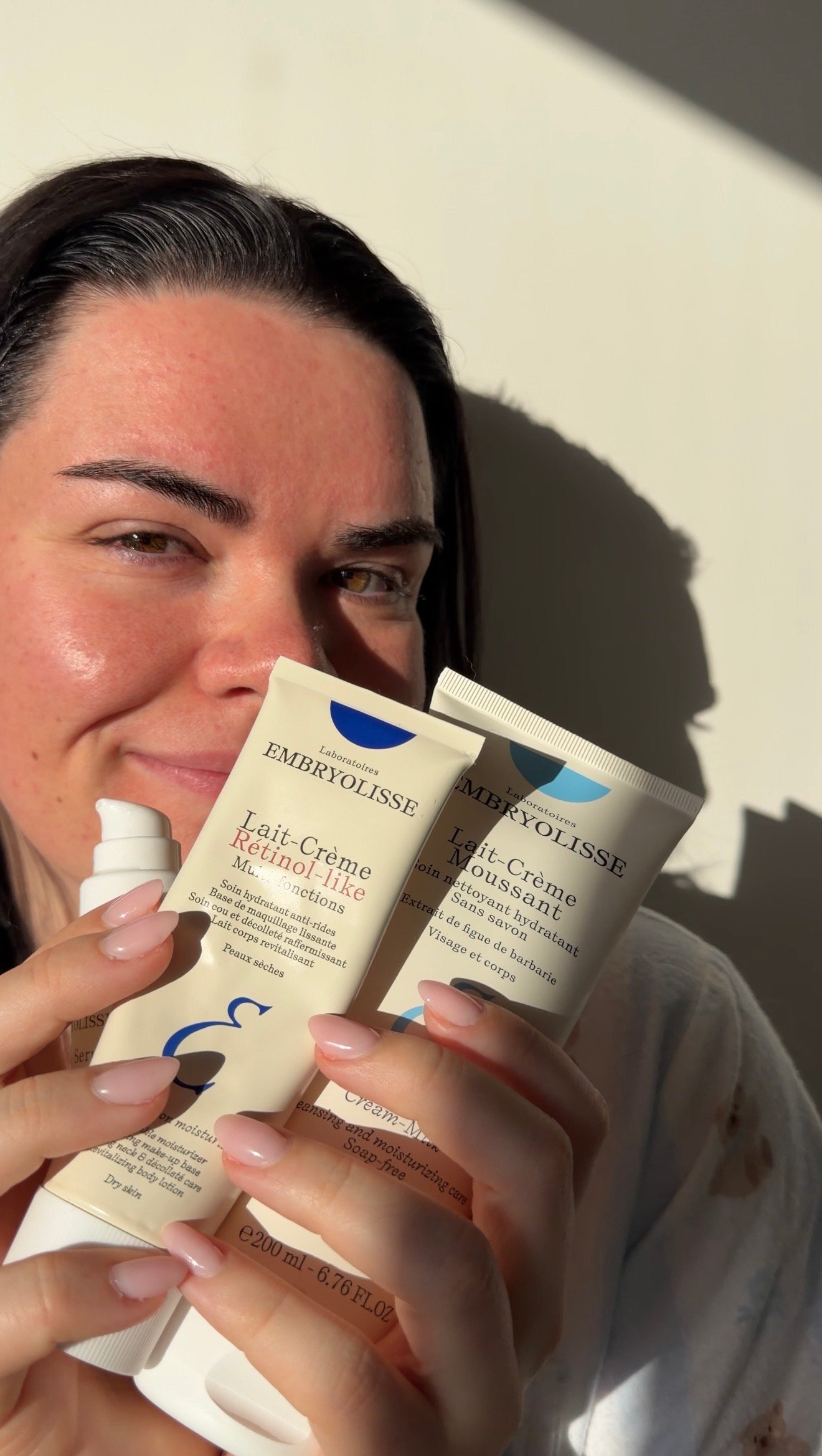 A woman holds two tubes of Embryolisse skincare products while smiling, with sunlight illuminating her face.