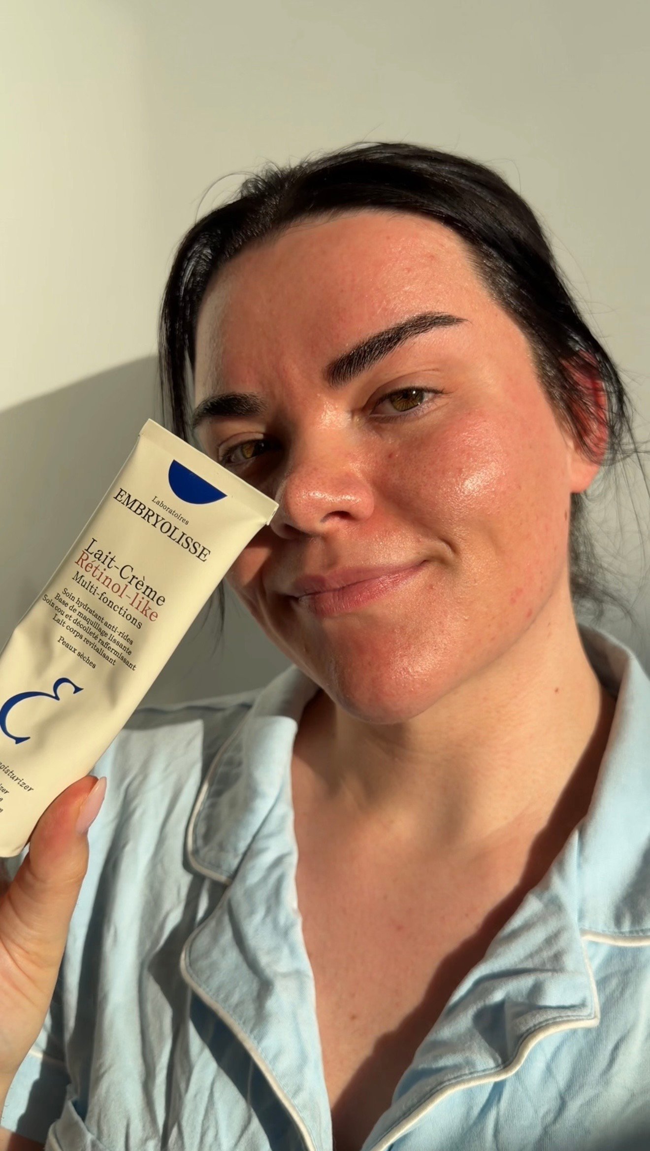 A person in light blue pajamas holds a tube of Embryolisse moisturizer, smiling softly at the camera.