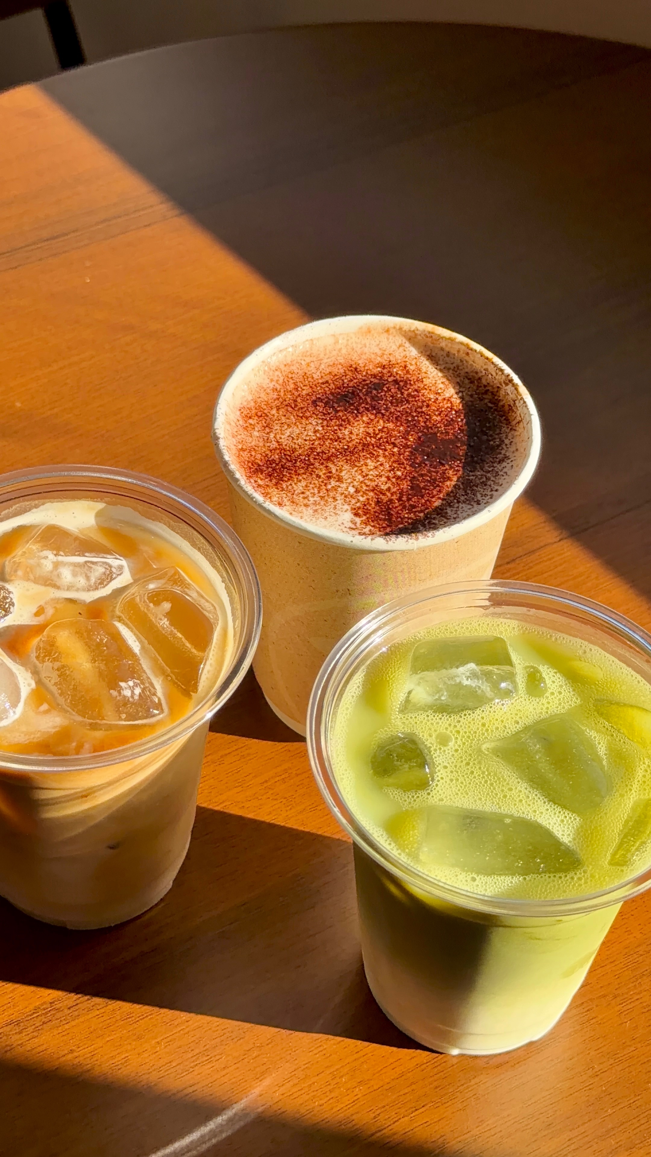 Three beverages on a wooden table: an iced coffee, a matcha drink, and a creamy drink topped with cinnamon.