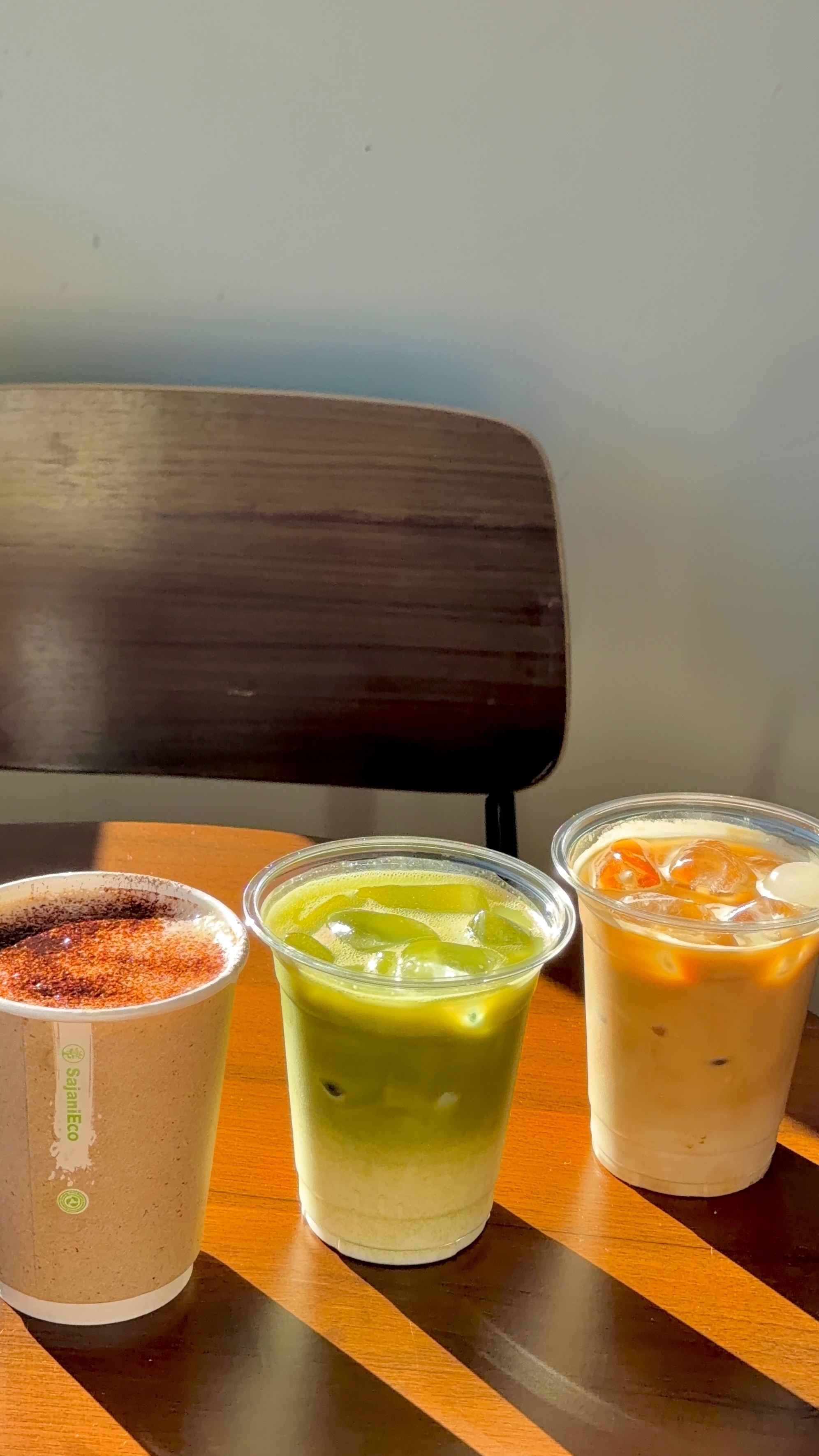 Three cups of different beverages on a wooden table, including a coffee, a matcha drink, and an iced tea.