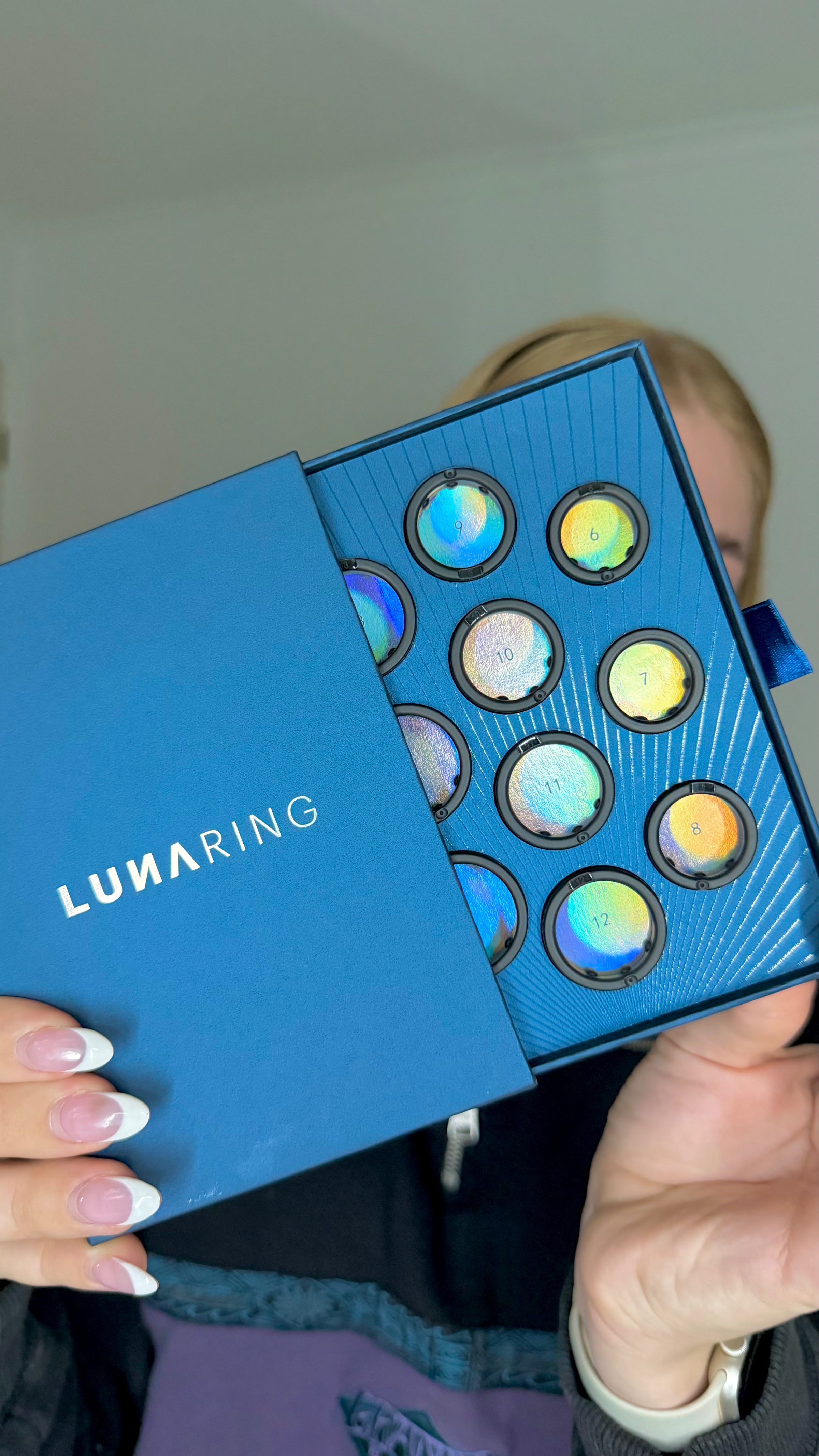 A person holding a blue box containing circular, iridescent items labeled 'LUNARING'. The background is softly blurred.