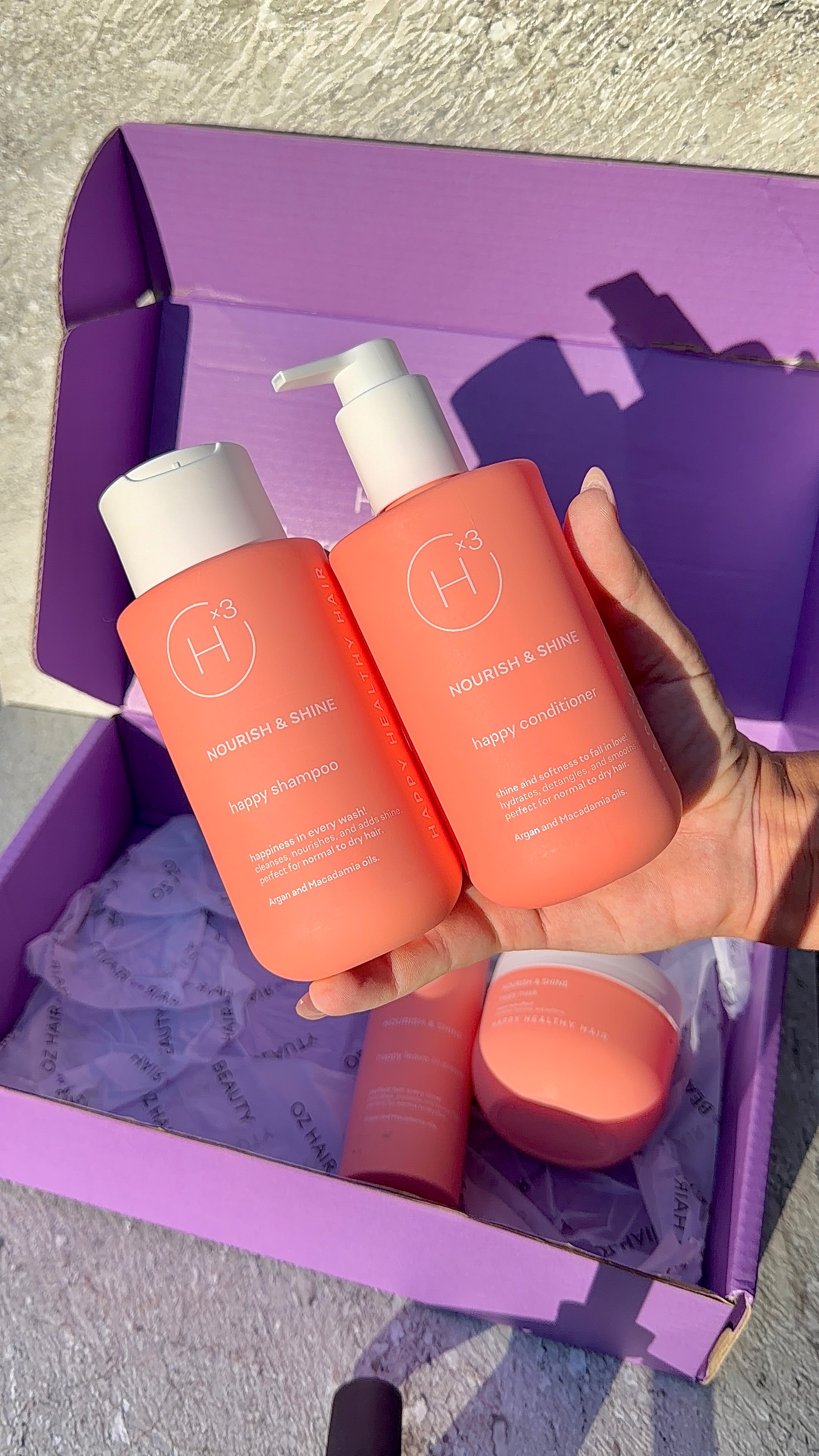 A hand holding two bottles of hair care products, labeled 'Nourish & Shine' and 'Happy Scalp Shampoo', in front of a purple box.