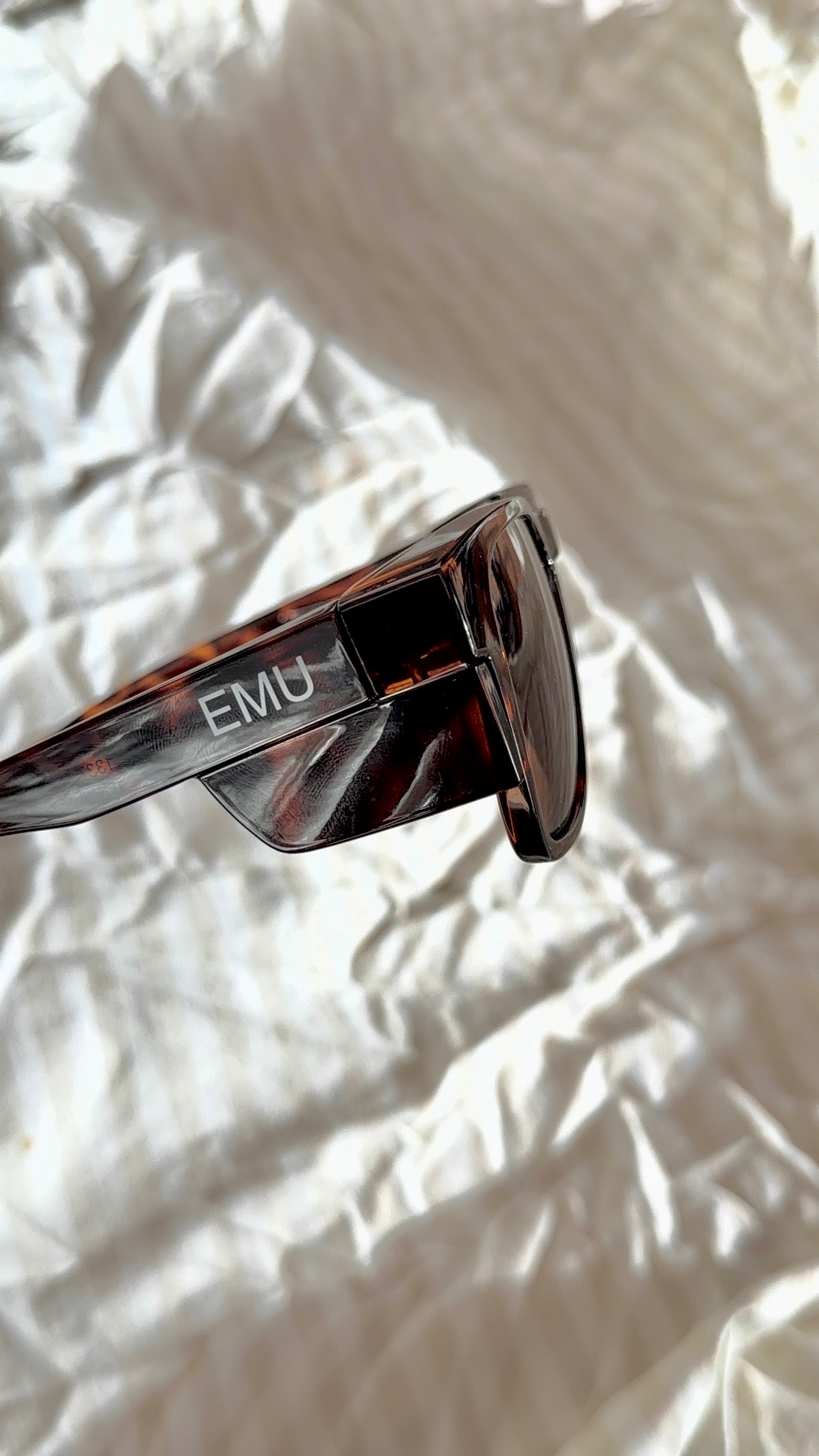 Close-up of a pair of stylish sunglasses with the brand name 'EMU' visible on the side, resting on a wrinkled white fabric.