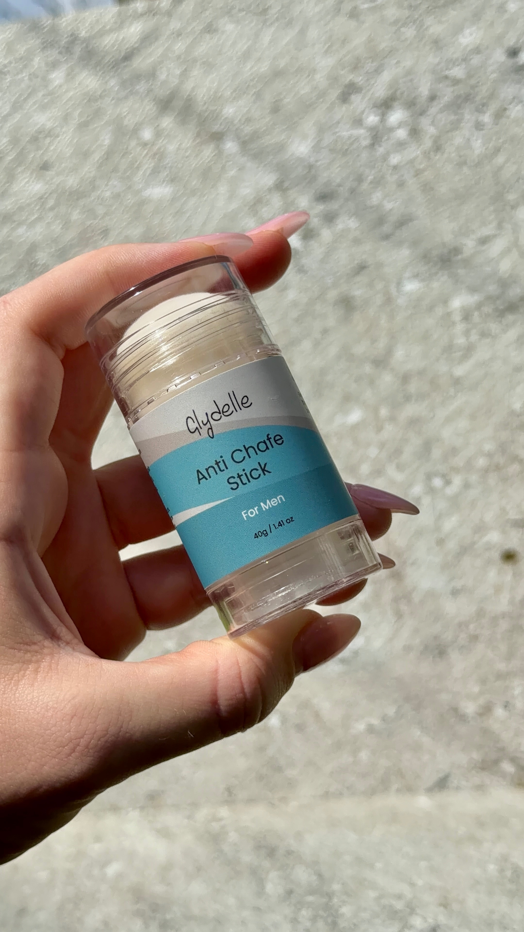 A hand holding a clear container of Clydeelle Anti Chafe Stick for men against a textured background.