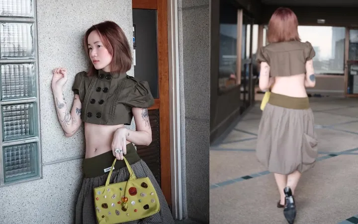 A person poses in a stylish cropped top and skirt, holding a yellow handbag adorned with colorful embellishments. The setting features a modern architectural background.